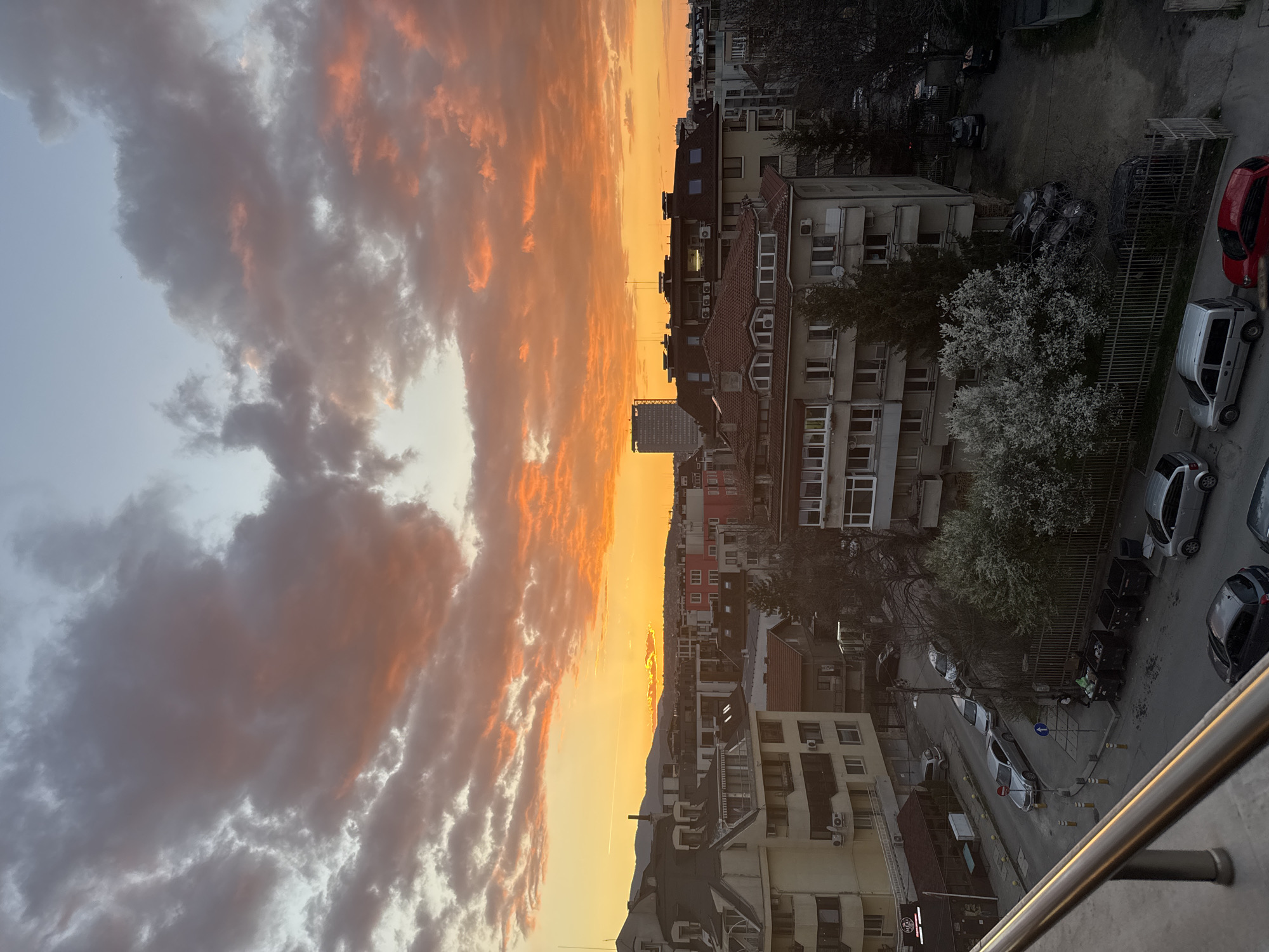 The image is a vertically-oriented shot taken from a high vantage point, likely a balcony. The foreground shows a metal railing, suggesting the vantage point is a balcony. The main subject is the sky, filled with clouds illuminated by a sunset, casting an orange and pink glow. Below the horizon line, there is a view of an urban landscape. Buildings of varying heights and architectural styles populate the scene, typical of Sofia, Bulgaria. Streets below are lined with parked cars. A tall building is visible in the distance. The image captures an urban sunset scene in Sofia, Bulgaria.