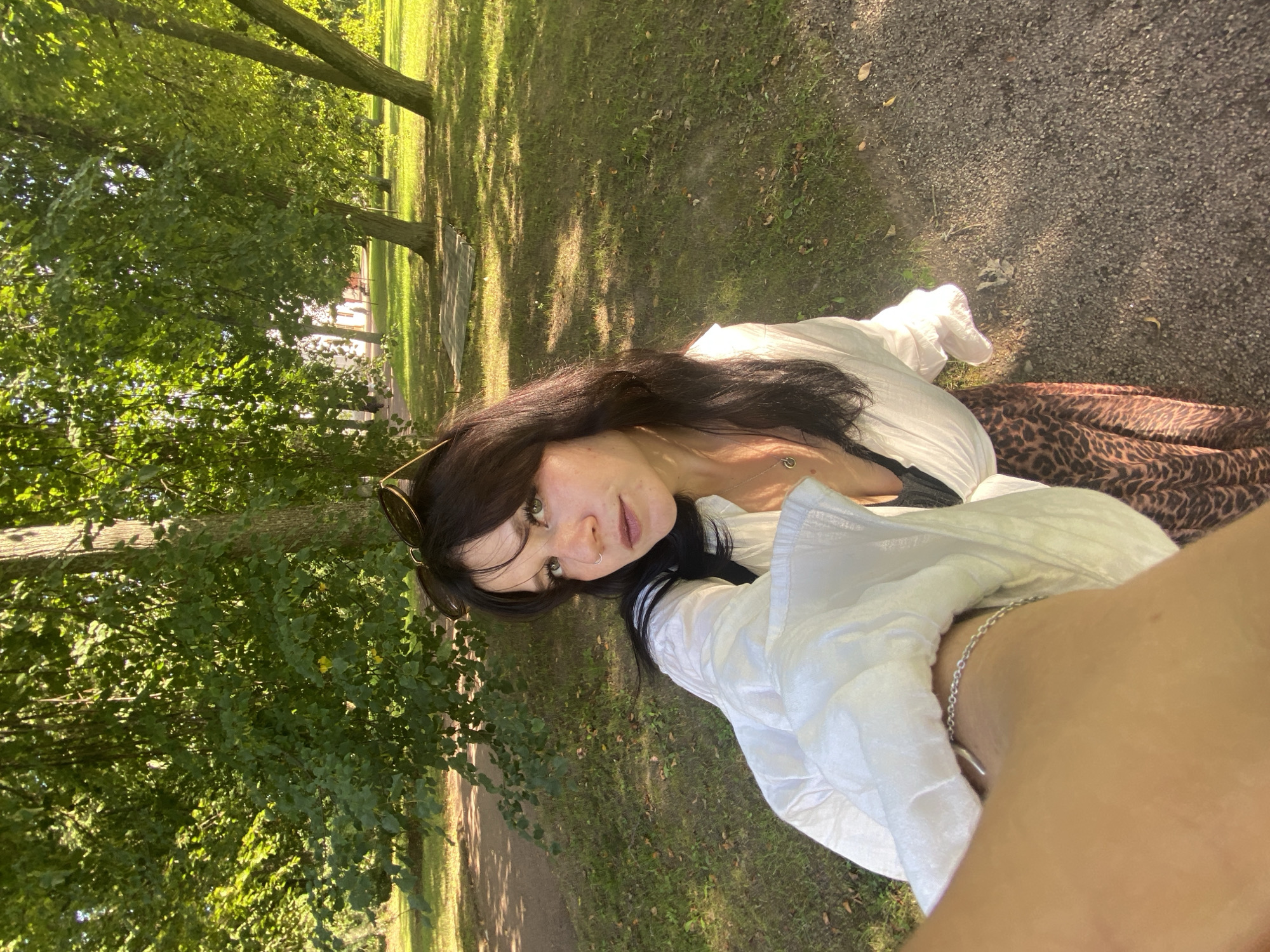 A young woman with dark hair and light eyes is taking a selfie outdoors in a park. She is wearing a white open shirt, a black top underneath, and a leopard print skirt. She has a silver bracelet on her wrist. Sunglasses are perched on her head. The background shows trees, grass, and a gravel path. The location appears to be the Lomonosov State Museum-Reserve, also known as Oranienbaum, in Lomonosov, Russia, given the architectural style visible in the distance and the park setting. The lighting suggests it is a sunny day.