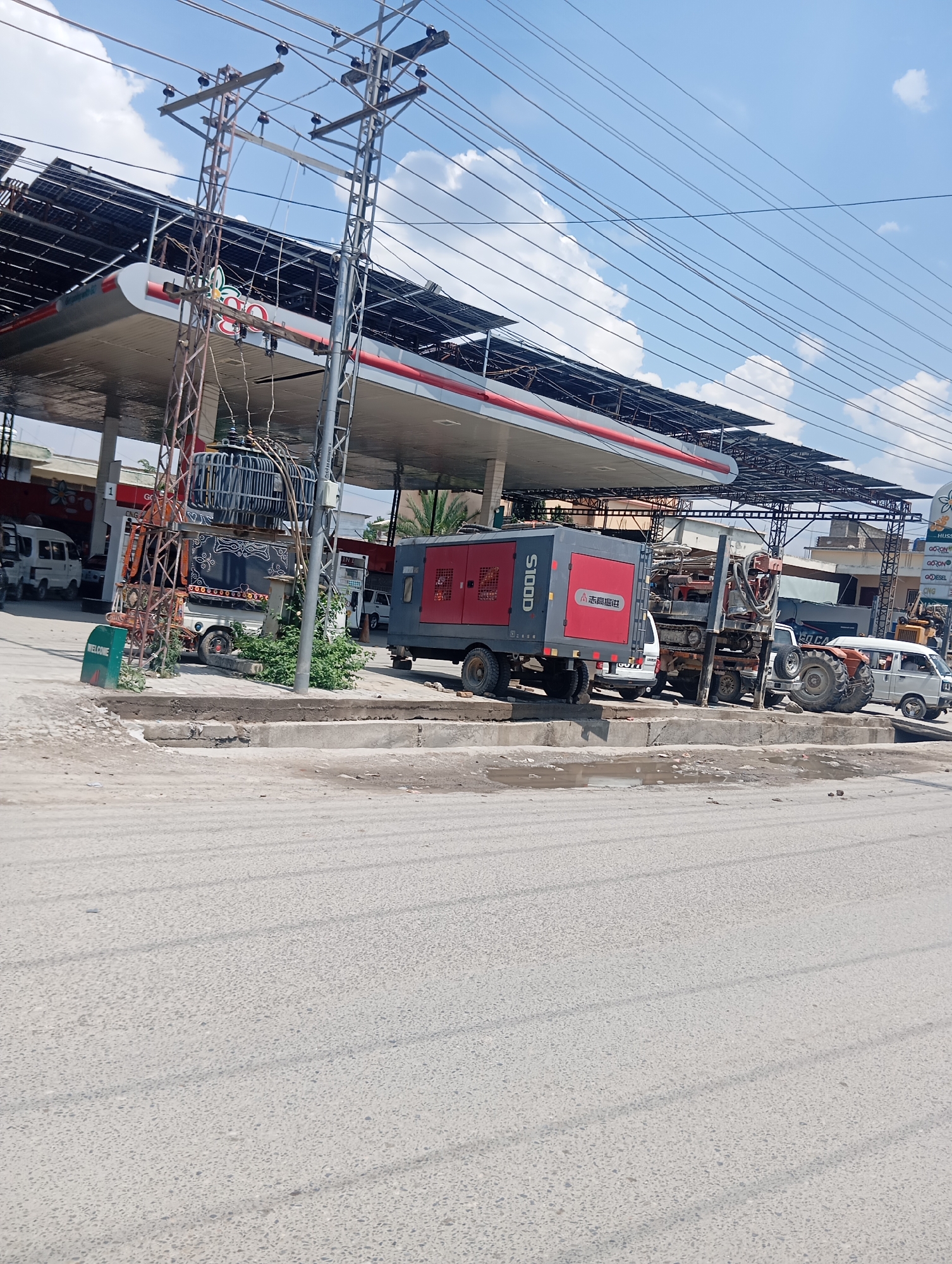 A gas station with solar panels on the roof is depicted under a cloudy sky. In the foreground, a gray and red air compressor is parked, with a drilling rig and a tractor visible behind it. Several white vans and a truck are also present at the station, along with a transformer. A green sign with the word "WELCOME" is positioned to the left of the image. The ground is paved with asphalt.