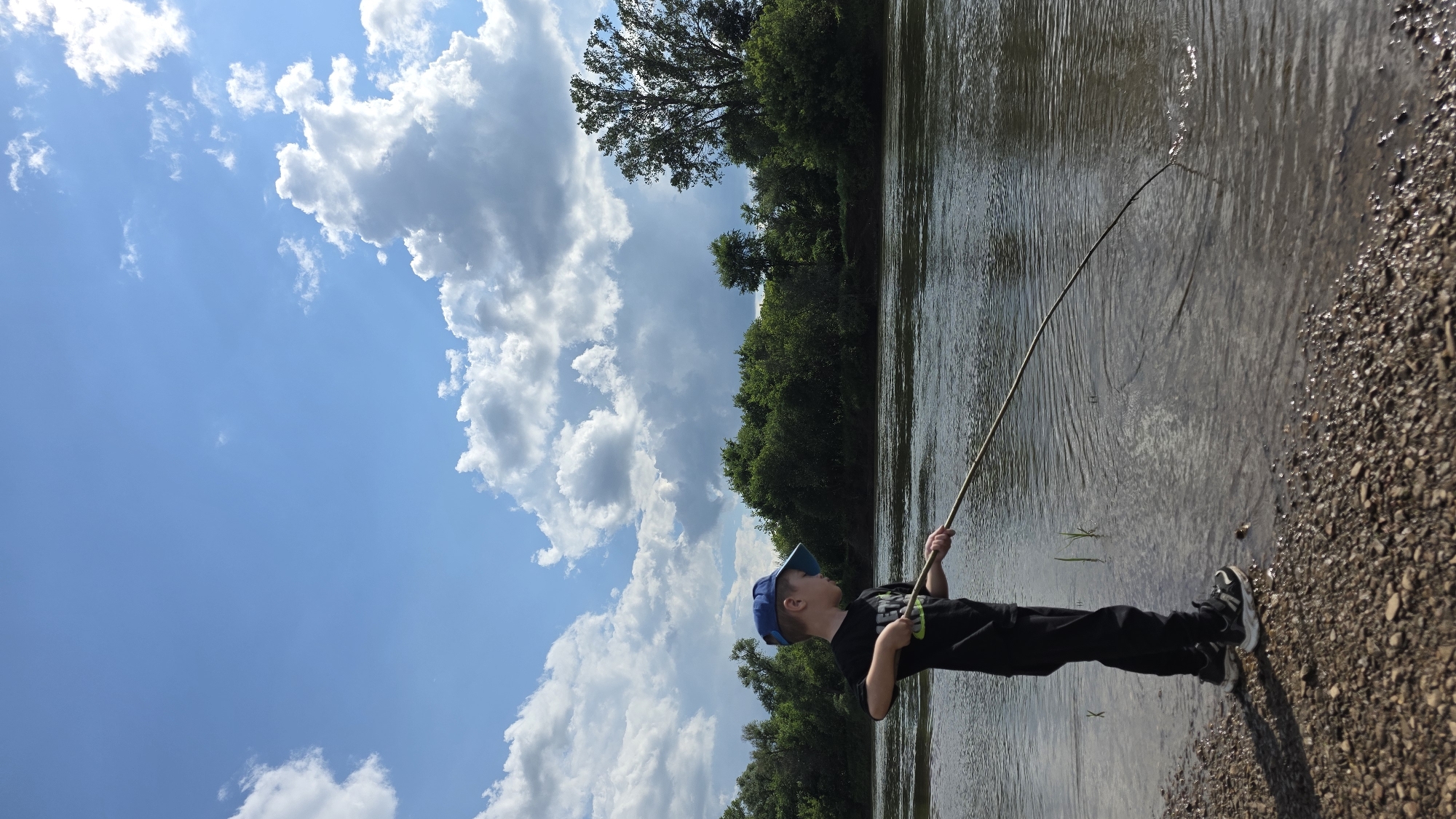 A young boy wearing a blue baseball cap, black t-shirt, and black pants is standing on the rocky shore of a river, holding a long stick which he is using to prod the water. The river is calm with ripples on the surface. Dense green trees line the opposite bank of the river, and a large tree with dark green leaves stands on the far right. The sky above is a vibrant blue with scattered white clouds, some of which are large and fluffy. The overall scene depicts a boy fishing or playing by the river on a bright, cloudy day.