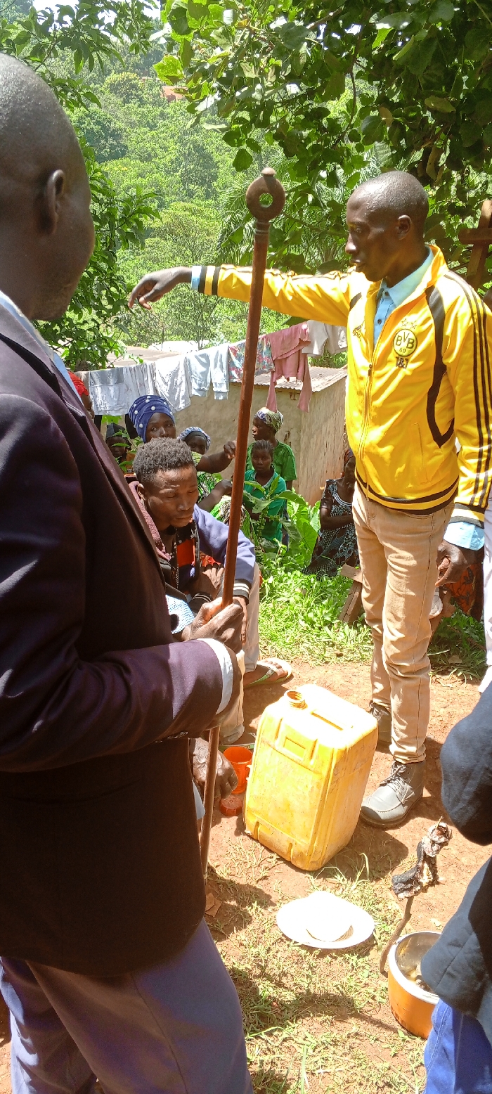 An outdoor gathering takes place in Kundu 1, Democratic Republic of the Congo, under bright sunlight and lush green foliage. In the foreground, partially visible from the back, is a man wearing a dark suit jacket, light blue shirt, and light blue trousers. He holds a long, brown wooden staff with a distinctive circular top. To his right, facing left, stands another man in a vibrant yellow jacket with black stripes (bearing "BVB 09" and "1&1" logos, indicating a Borussia Dortmund football club jacket), a light blue collared shirt, and light brown trousers. He is actively gesturing or pointing with his right hand towards an unseen area to the left.
Seated below him and to the left, a younger man with dark, braided hair wears a dark blue/grey hooded top and looks downwards. Behind these central figures, a group of women and children are gathered, some partially obscured by vegetation. One woman in a blue head covering looks directly at the camera. In the mid-ground, a clothesline with various items of laundry hangs between simple, light-colored structures with corrugated iron roofs.
On the dusty ground, near the feet of the standing man, rests a yellow plastic jerrycan, open at the top. Beside it are two white plates and an orange pot or container holding dark contents. The scene suggests a community meeting or traditional gathering, with the staffs possibly indicating a ceremonial or authoritative role for some participants. The natural environment is dominated by dense trees and greenery, hinting at a rural or forested location.