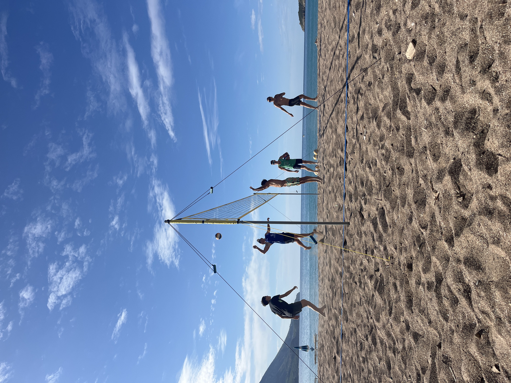 The image shows a group of six men playing volleyball on a beach in Bečići, Montenegro. The scene is composed of a volleyball net, a sand beach, a blue sky with clouds, and the Adriatic Sea in the background. The men are positioned at different points of the net, in various athletic poses, suggesting they are actively participating in a volleyball game. The beach itself has dark-colored sand. A mountain slope appears in the lower left corner of the image, offering additional context for the geographical location.