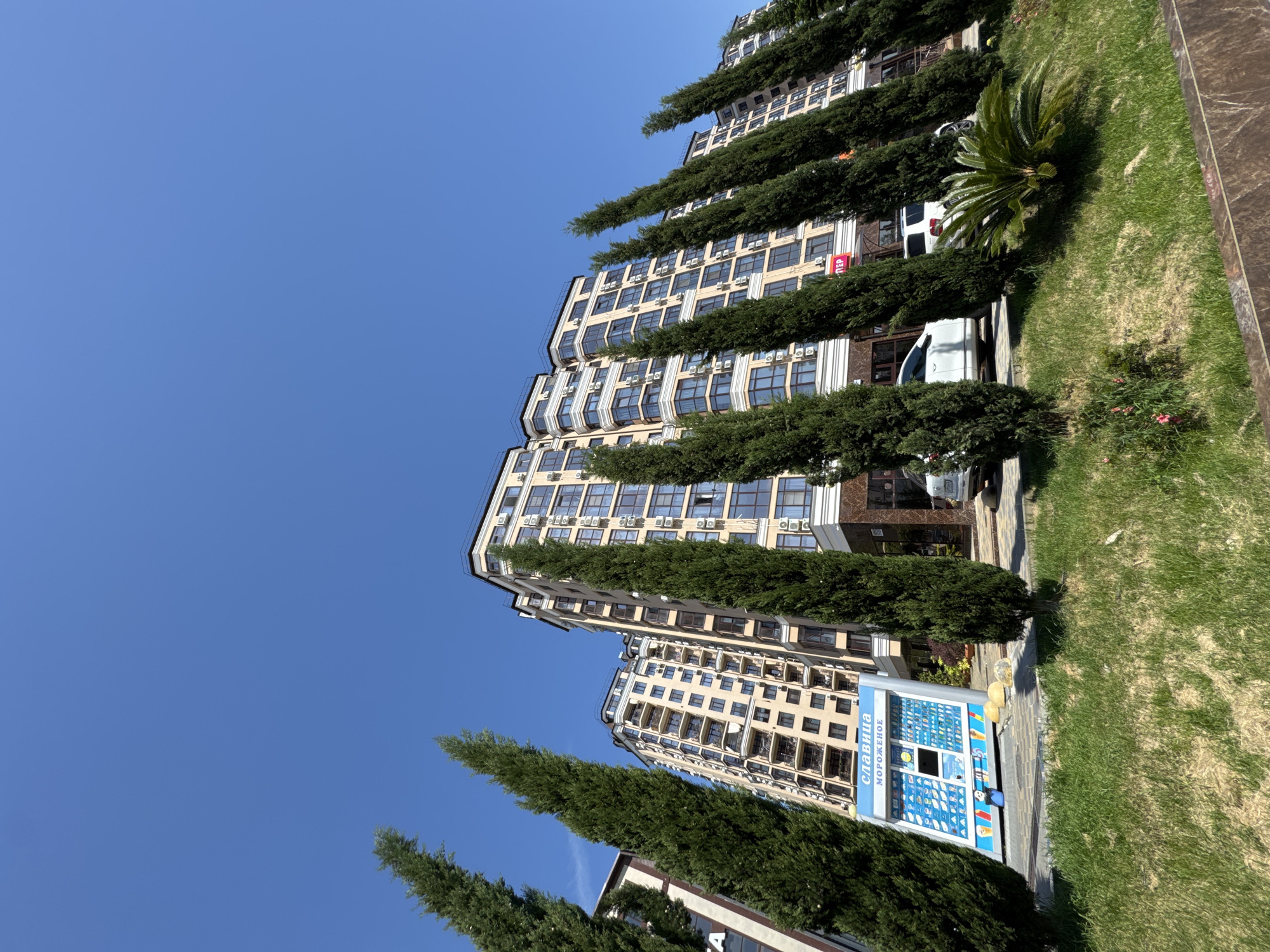 The image captures an outdoor urban scene under a bright, clear blue sky, suggesting daytime. The primary subjects are several modern multi-story buildings, characterized by their light-colored facades, numerous windows, and balconies. Many air conditioning units are visible on the exterior walls of the buildings.
Rows of tall, slender, dark green coniferous trees, possibly cypresses, are prominent in the foreground and midground, partially obscuring the buildings. To the right of the trees, there is a manicured grassy area with some shrubs and a distinct palm-like plant with spiky leaves. A paved street or sidewalk runs parallel to the buildings and trees.
A white van and part of another white car are visible parked between the trees. In the lower right section of the image, a blue and white vending machine stands on the sidewalk. Visible text on the machine is in Cyrillic: "СЛАВИЦА" (Slavitsa) and "МОРОЖЕНОЕ" (Morozhenoe), indicating it is an ice cream vending machine. A small red sign with illegible white text can also be seen on one of the buildings.
No people are visible, suggesting a quiet street or residential area. The scene depicts a sunny day with no clouds, reflecting the pleasant weather of Sochi, Russia. The combination of modern architecture and lush greenery creates an appealing urban landscape.