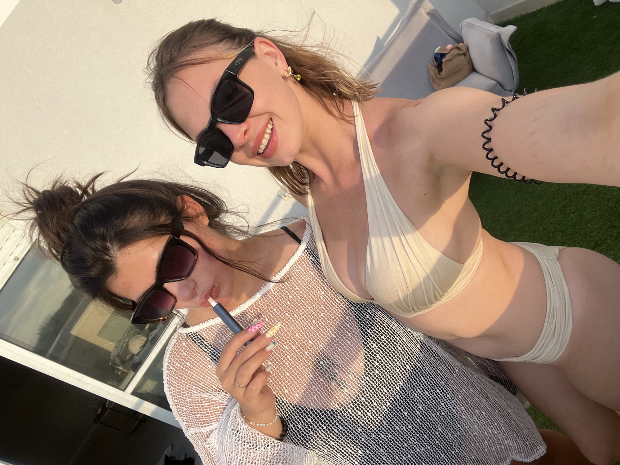 Two young women are photographed in what appears to be an outdoor setting, possibly a balcony or rooftop, with a white wall in the background. The woman on the left, with dark hair pulled into a bun, wears a white mesh cover-up over a dark bikini top and sunglasses. She is holding a device to her mouth, resembling an e-cigarette. Her nails are painted in various bright colors and patterns. The woman on the right, with lighter brown hair, is wearing a beige bikini and sunglasses. She is smiling and has a decorative earring in her left ear. The lighting suggests it is late afternoon or early evening, with warm sunlight casting shadows. There are some cushions and a woven bag visible in the background, further indicating a leisure or outdoor living space. There is no indication of the specific location being the city of Ashdod, Israel.