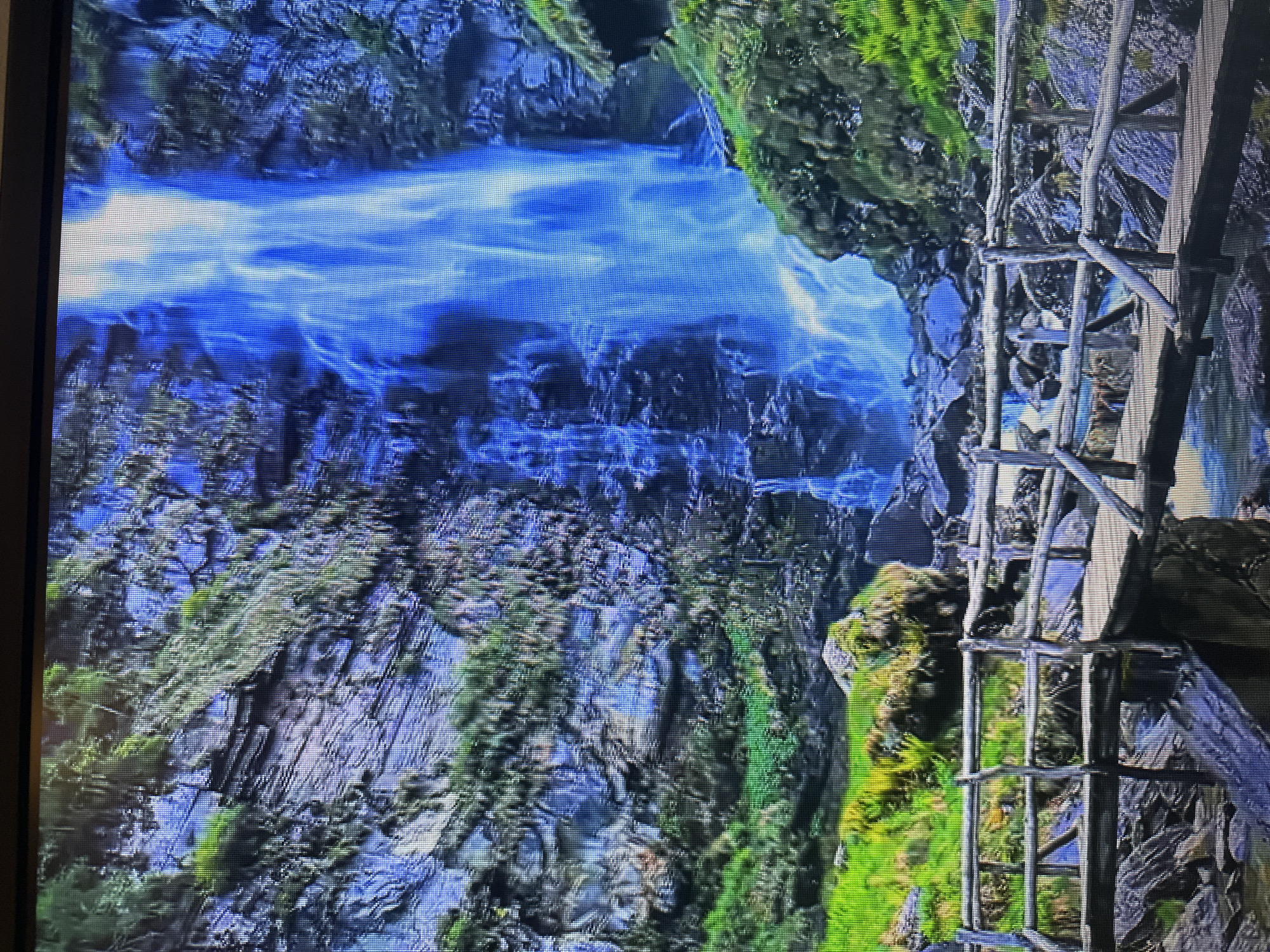 The image depicts a powerful waterfall cascading down a rocky cliff face. The water is a brilliant blue, with streaks of white where it crashes and churns. Lush green vegetation covers parts of the cliff and the surrounding landscape. In the foreground, a rustic wooden bridge crosses over a stream, its surface weathered and built from natural logs and branches. The bridge leads towards the waterfall, offering a vantage point to admire the natural spectacle. The overall scene is one of wild, untamed beauty. This image does not depict any location in Kyiv, Ukraine.