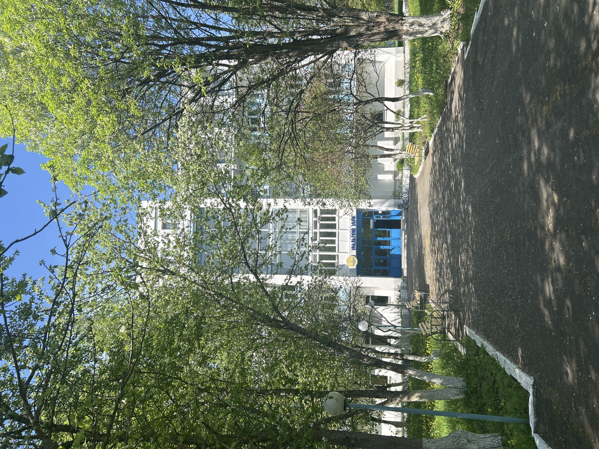 The image depicts a multi-story building in Noviy Urengoy, Russia. The building has a white facade with multiple windows and a blue entrance area. Trees with green leaves partially obscure the view of the building. A paved pathway leads towards the entrance, and there are benches and painted tree trunks along the side of the pathway. The sky is visible in the upper left corner.
