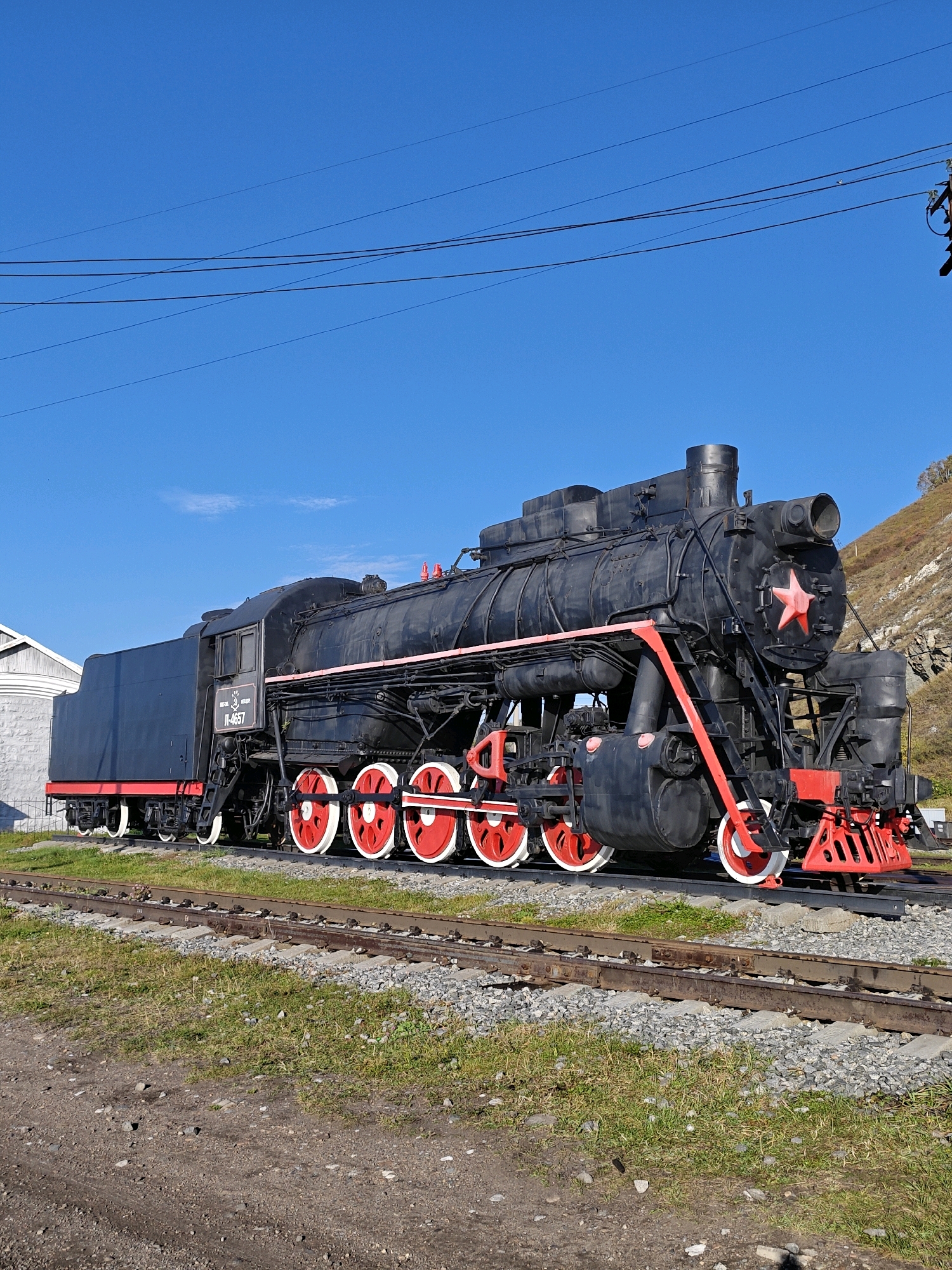 This is a wide, eye-level shot of a black steam locomotive on railroad tracks in Listvyanka, Russia, under a clear blue sky. The locomotive is the primary subject, detailed with red accents on its wheels, trim, and a prominent red star on the front. The train is stationary on a double set of tracks surrounded by gravel and green grass. To the left, a white building with a grey stone base is partially visible. In the background, a steep, rocky hillside slopes upwards. Overhead, power lines crisscross the sky, and a small bird is perched on one of them. The overall scene depicts an outdoor display or museum setting in the town of Listvyanka.