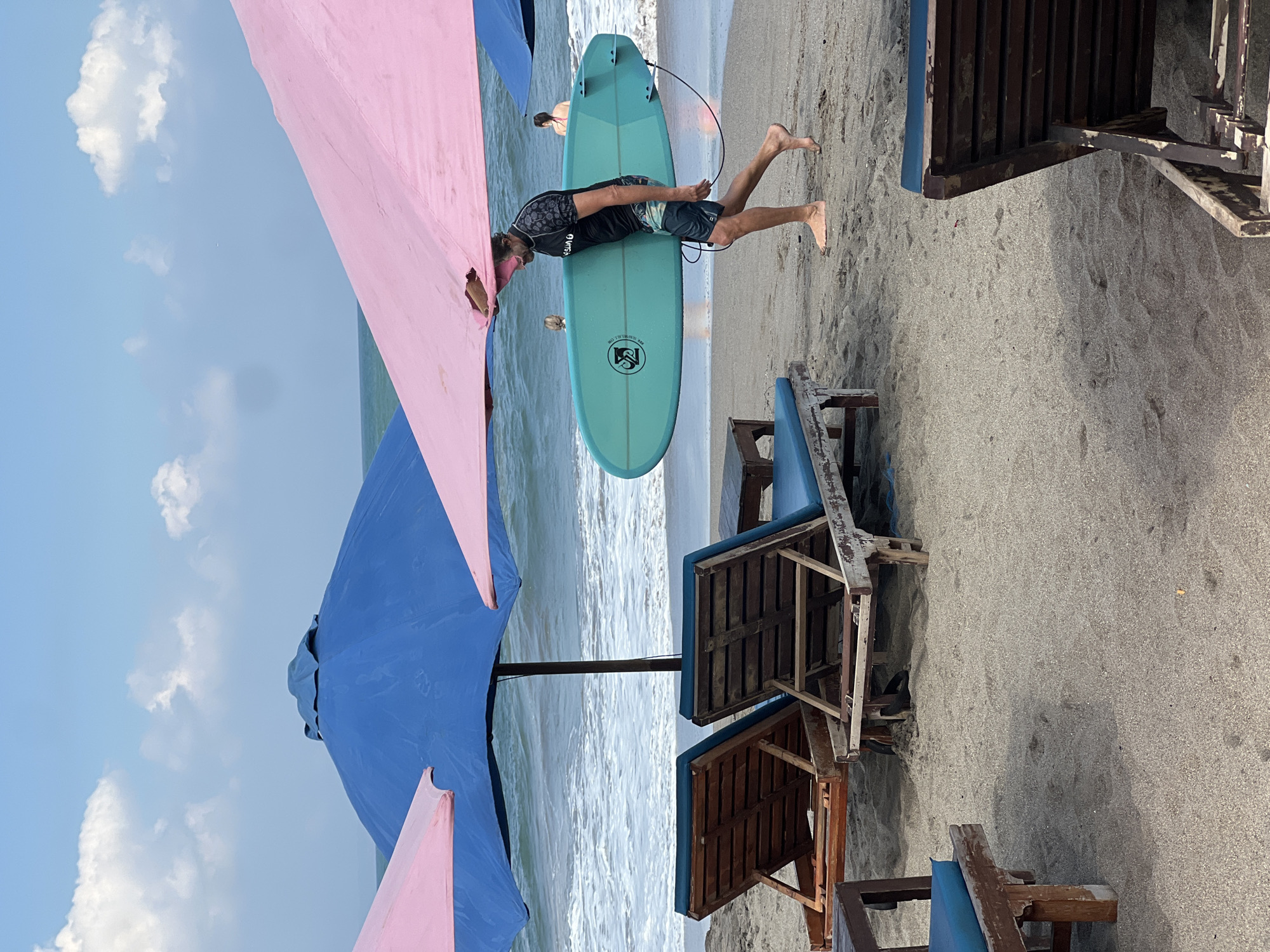 The image depicts a beachfront scene in North Kuta, Indonesia, characterized by a sandy shore and the ocean. In the foreground, several weathered wooden lounge chairs with blue cushions are arranged on the sand. A large pink umbrella partially shades the area. Dominating the mid-ground is a man walking along the beach, carrying a turquoise surfboard under his right arm. He is wearing a black rash guard and patterned shorts. His left hand is holding the top edge of the pink umbrella. In the background, the ocean waves are visible, and two other individuals, a woman and a child, are present near the water's edge. A large blue umbrella is also visible in the scene, positioned behind the pink one. The sky is partly cloudy.