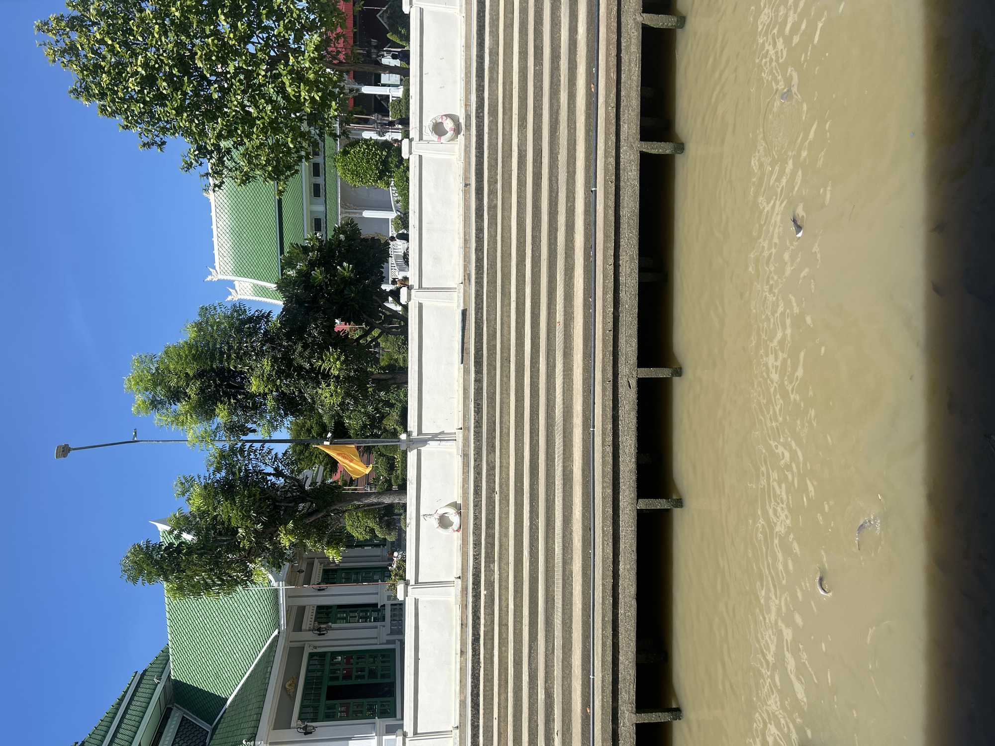 This is a medium shot of a canal in Thai Ko Subdistrict, Thailand, on a sunny day. The canal water is a murky brown color. Several fish are visible swimming just below the surface. The canal is bordered by a concrete embankment with a series of wide steps leading up to a white concrete wall. On the wall, a life preserver is hanging. Behind the wall is a building with a green tiled roof and white walls, featuring green doors and windows. Trees with lush green foliage are situated around the building and behind the wall. A yellow flag is flying from a pole. In the distance, several people can be seen walking along the walkway behind the wall.