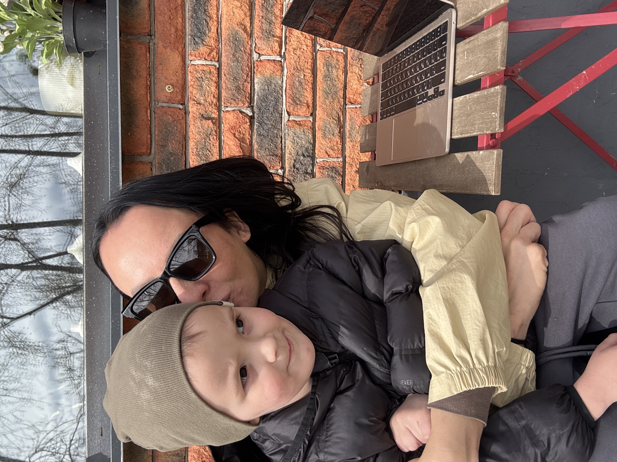 An upright outdoor shot captures an adult embracing a young child in Chaikovsky, Russia. The adult, on the left, has long dark hair and wears black sunglasses, with their head leaning affectionately towards the child. They are dressed in a light-colored, possibly beige, lightweight jacket. The young child, held closely by the adult, wears a grey-green beanie and a black puffer jacket, looking directly at the viewer with a mild expression.
To the right and slightly behind the subjects, a silver laptop rests on a rustic outdoor table or bench, which features wooden slats and a bright red metal frame. A warm-toned brick wall serves as the background behind the table and part of the subjects. On the far left, a window or glass pane reflects the intricate, bare branches of trees against a bright, clear sky, suggesting sunny weather and a natural outdoor environment. A potted plant is also visible in the upper left corner.
The bright, direct lighting indicates daytime, likely morning or afternoon. The bare trees in the reflection suggest the season is late autumn, winter, or early spring. The child's black puffer jacket has the word "EVER" faintly visible on its lower sleeve. The image portrays a tender moment of affection and companionship between the adult and child in an outdoor setting.