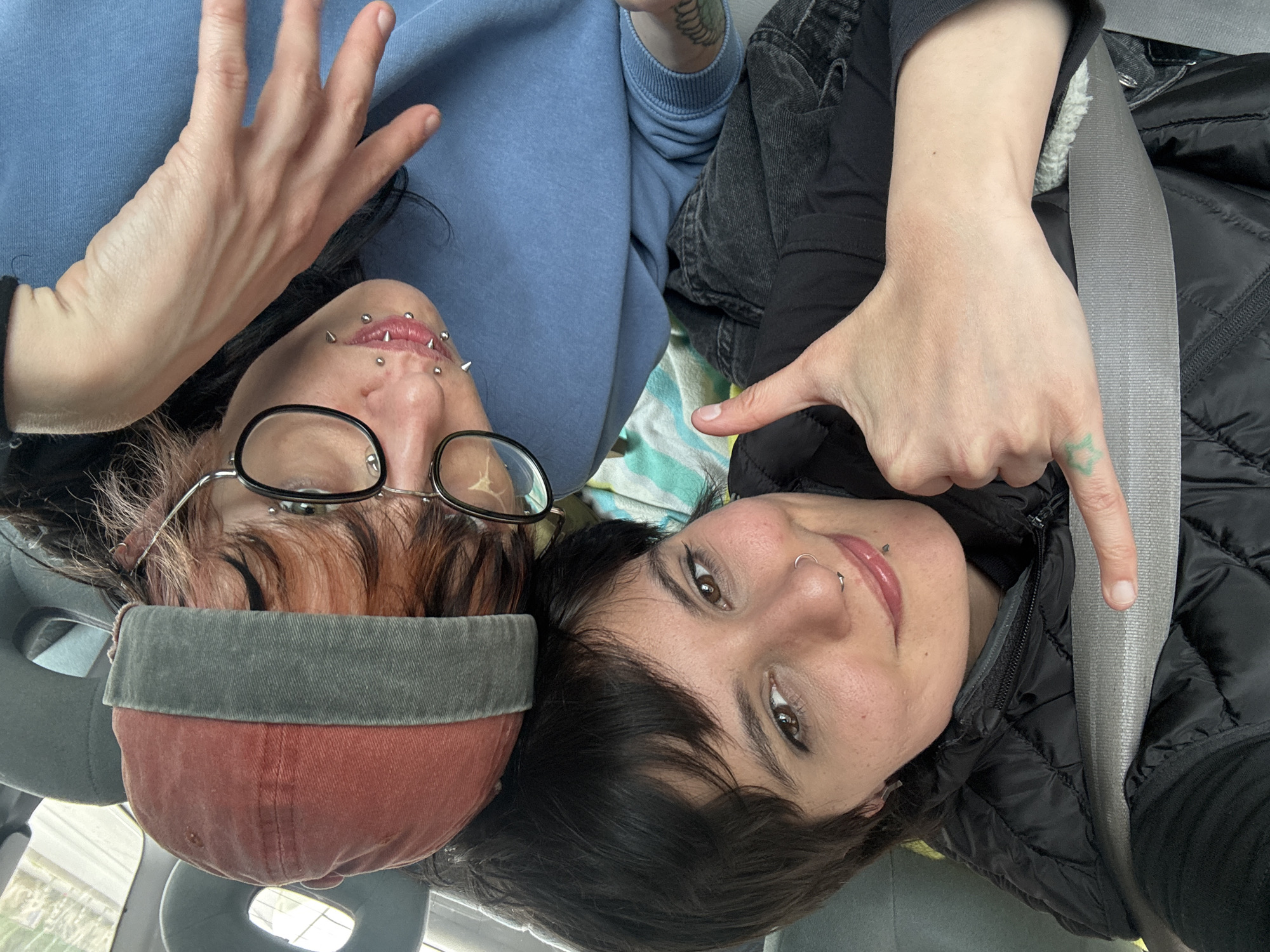 Two individuals are captured in what appears to be a selfie taken from an overhead perspective, inside a vehicle in Sofia, Bulgaria. The image shows their heads and upper bodies, oriented upside down.
On the left, one person is wearing a light blue long-sleeved top or hoodie and a red and grey baseball cap, pulled low over their forehead. They have dark hair, possibly with reddish highlights, and wear black-rimmed glasses. Their face features multiple piercings, including prominent spiked lip piercings, a philtrum piercing, and a bridge piercing. Their right hand is raised, with the palm facing the camera and fingers splayed open.
Next to them, on the right, the second person is visible. They are wearing a black puffer jacket and dark jeans, secured by a grey car seatbelt that runs diagonally across their chest. This individual has short dark hair and a septum piercing. Their left hand is visible, pointing downwards, with a small green star-shaped tattoo on the back of their hand.
Both individuals have neutral to slightly smiling expressions, appearing relaxed. Parts of the car's interior, including seat headrests and a patterned blanket or fabric with green, white, and blue stripes, are visible in the background. There is no visible text in the image.