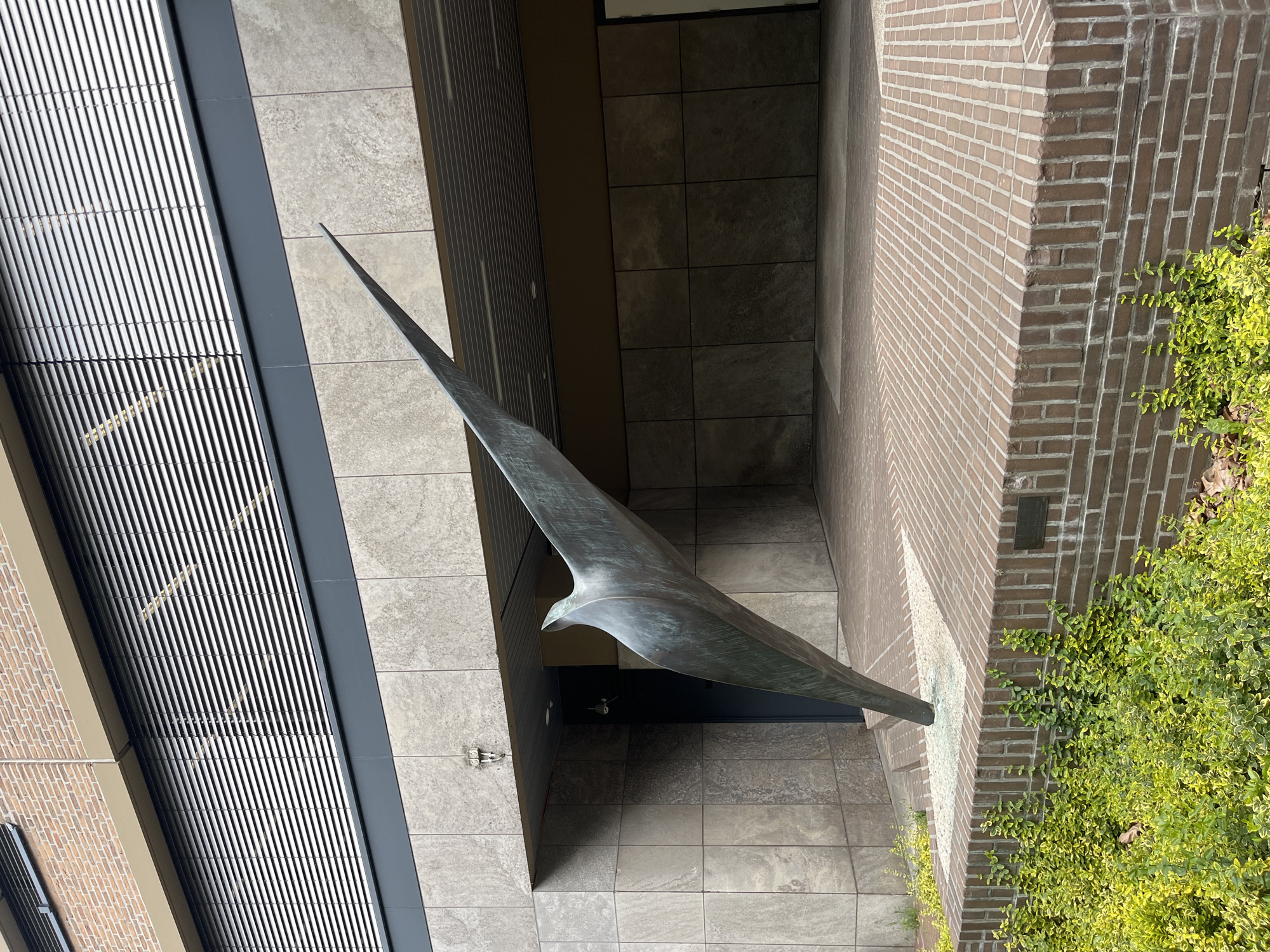 A bronze sculpture of a bird in flight is positioned in front of a building with a brick facade and a section of horizontal blinds. The sculpture, which appears abstract and streamlined, extends from a base made of brick and white gravel. The building features large, square stone tiles on its exterior walls and a dark doorway to the left of the sculpture. Lush green bushes grow in front of the brick base. The ground is paved with brown bricks arranged in a herringbone pattern. The sky is not visible in the image.