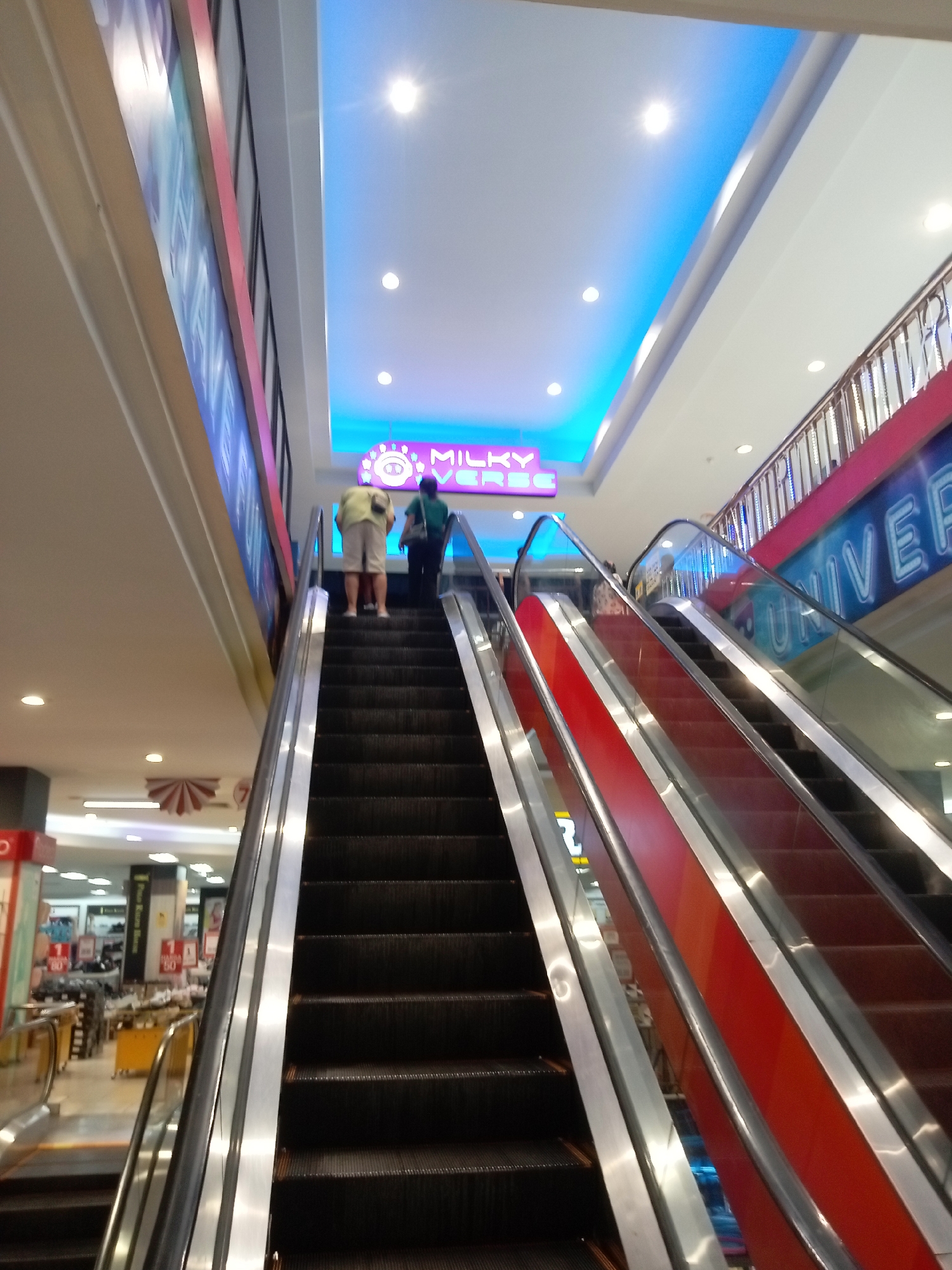 An interior shot of a shopping mall in Batam, Indonesia shows two escalators. The escalator on the left is dark gray with silver handrails. The escalator on the right is red with silver handrails. Two individuals are ascending the left escalator. The first individual, from behind, wears a light yellow short-sleeved shirt and light-colored shorts. The second individual, also from behind, wears a dark green short-sleeved shirt and dark pants. They are standing on the escalator steps, with the first individual on the left and the second on the right. The background features a brightly lit ceiling with blue accents and several circular light fixtures. A neon sign with purple lettering, possibly for "MILKY UNIVERSE," is visible above the escalators. The mall also appears to have multiple levels, with railings and architectural elements suggesting different floors. Storefronts and displays are visible in the blurred background. The lighting is bright, with some reflections on the escalator handrails and glass barriers.