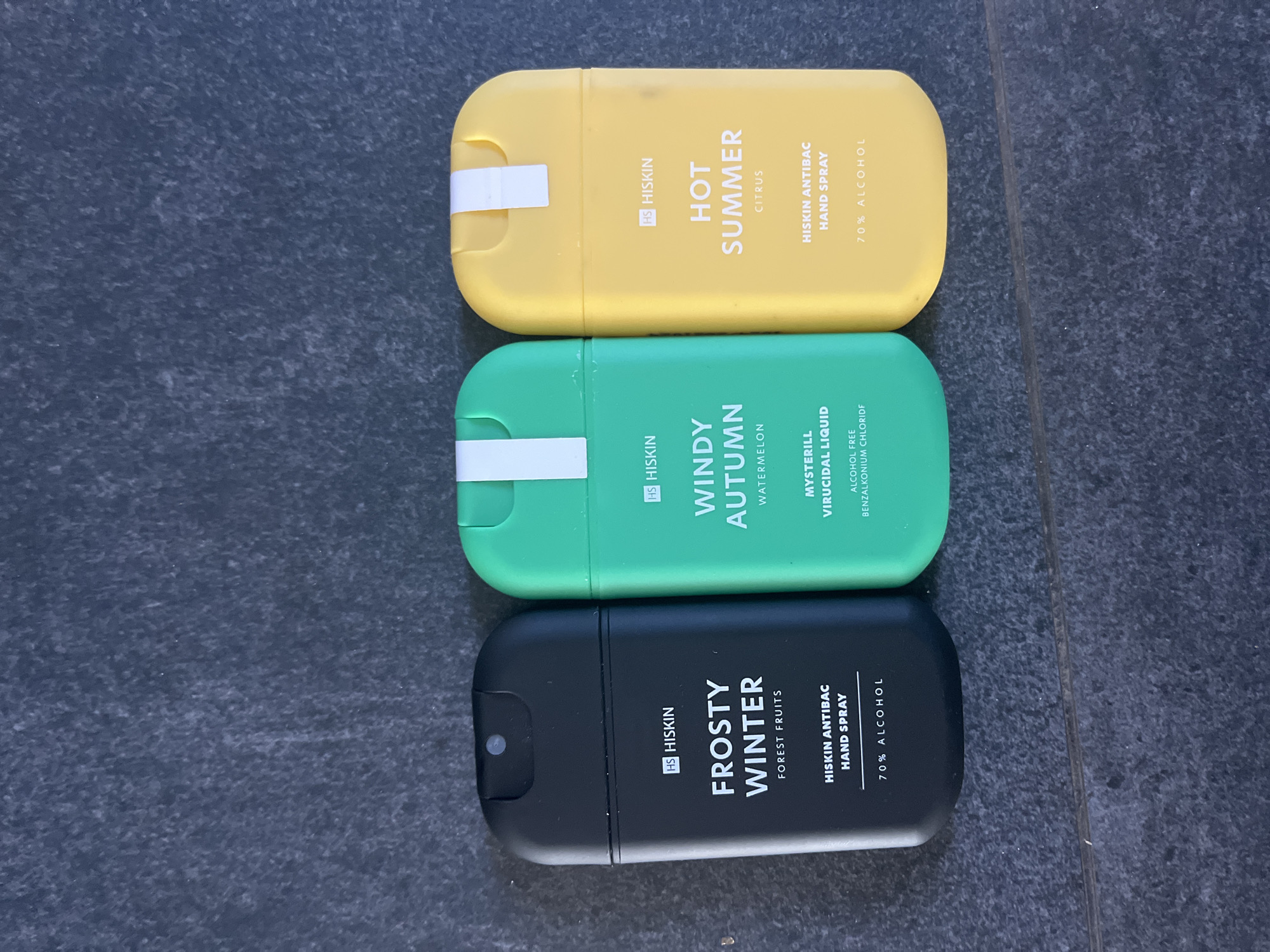 Three spray bottles of hand sanitizer are arranged on a dark gray tiled surface. From left to right, the bottles are black, green, and yellow. The black bottle has "HISKIN FROSTY WINTER FOREST FRUITS HISKIN ANTIBAC HAND SPRAY 70% ALCOHOL" printed on it. The green bottle has "HISKIN WINDY AUTUMN WATERMELON MYSTERILL VIRUCIDAL LIQUID ALCOHOL FREE BENZALKONIUM CHLORIDE" printed on it. The yellow bottle has "HISKIN HOT SUMMER CITRUS HISKIN ANTIBAC HAND SPRAY 70% ALCOHOL" printed on it. The surface is a dark gray, textured tile, likely a floor or countertop. The lighting suggests an indoor environment, possibly a retail or home setting. The image does not contain any elements that would indicate its location in Haabneeme alevik, Estonia.