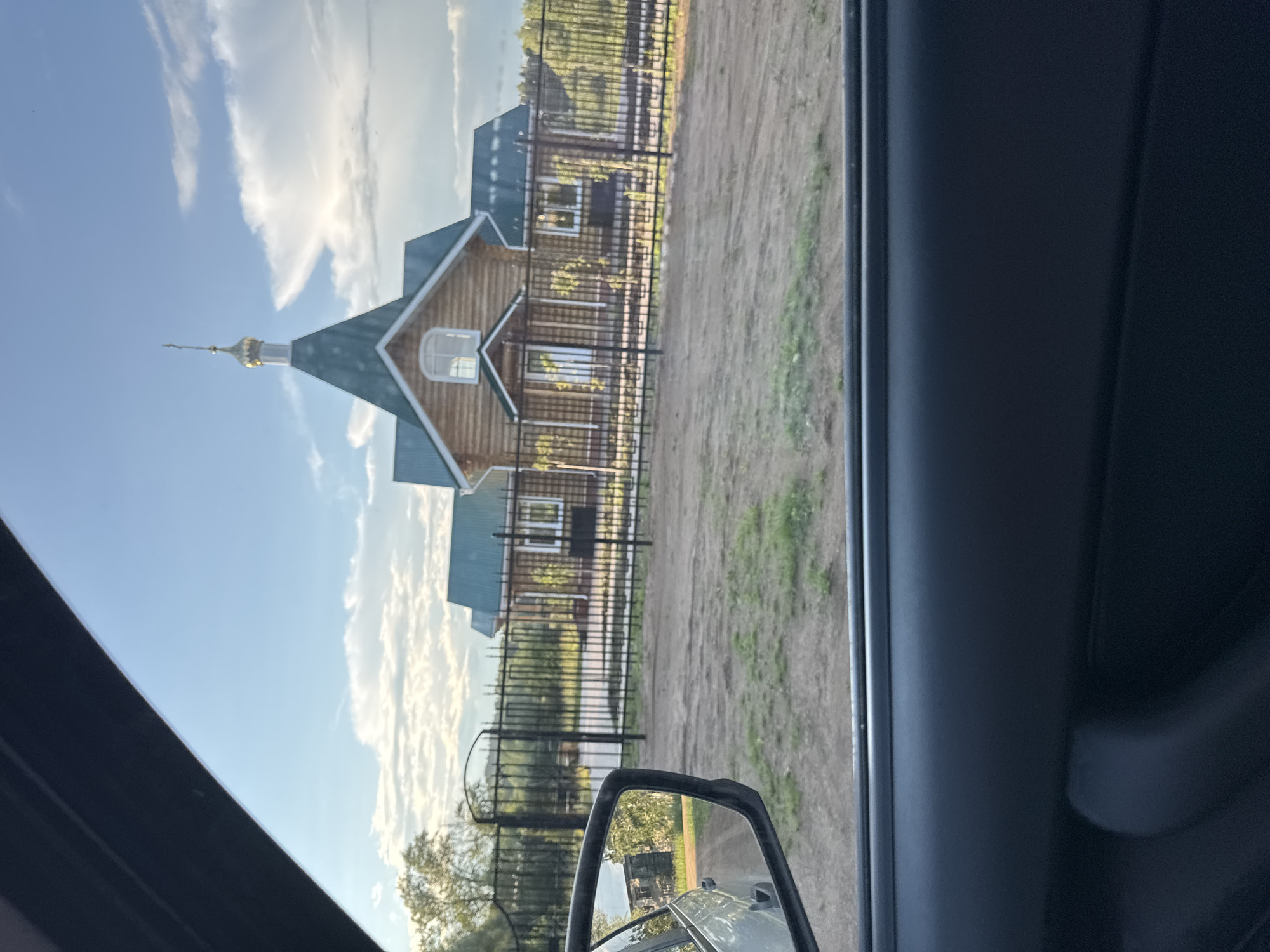 A wooden church with a green roof and a golden dome is visible through a car window in Вассята, Russia. The church is behind a black metal fence. Trees and a cloudy sky are in the background. The car's side mirror reflects a dirt road and trees. The interior of the car is also visible, including the door panel and the bottom of the window frame.