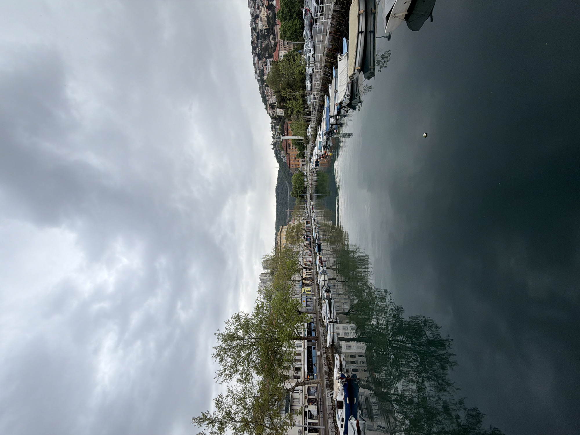 The image depicts the Mrtvi Kanal (Dead Channel) in the city of Grad Rijeka, Croatia. The scene is taken from street level, looking across the water towards the opposite bank. Rows of buildings and trees line the waterfront, and several boats are docked alongside the quayside. The water reflects the buildings and cloudy sky above. There are no visible people performing discernible actions in the scene.