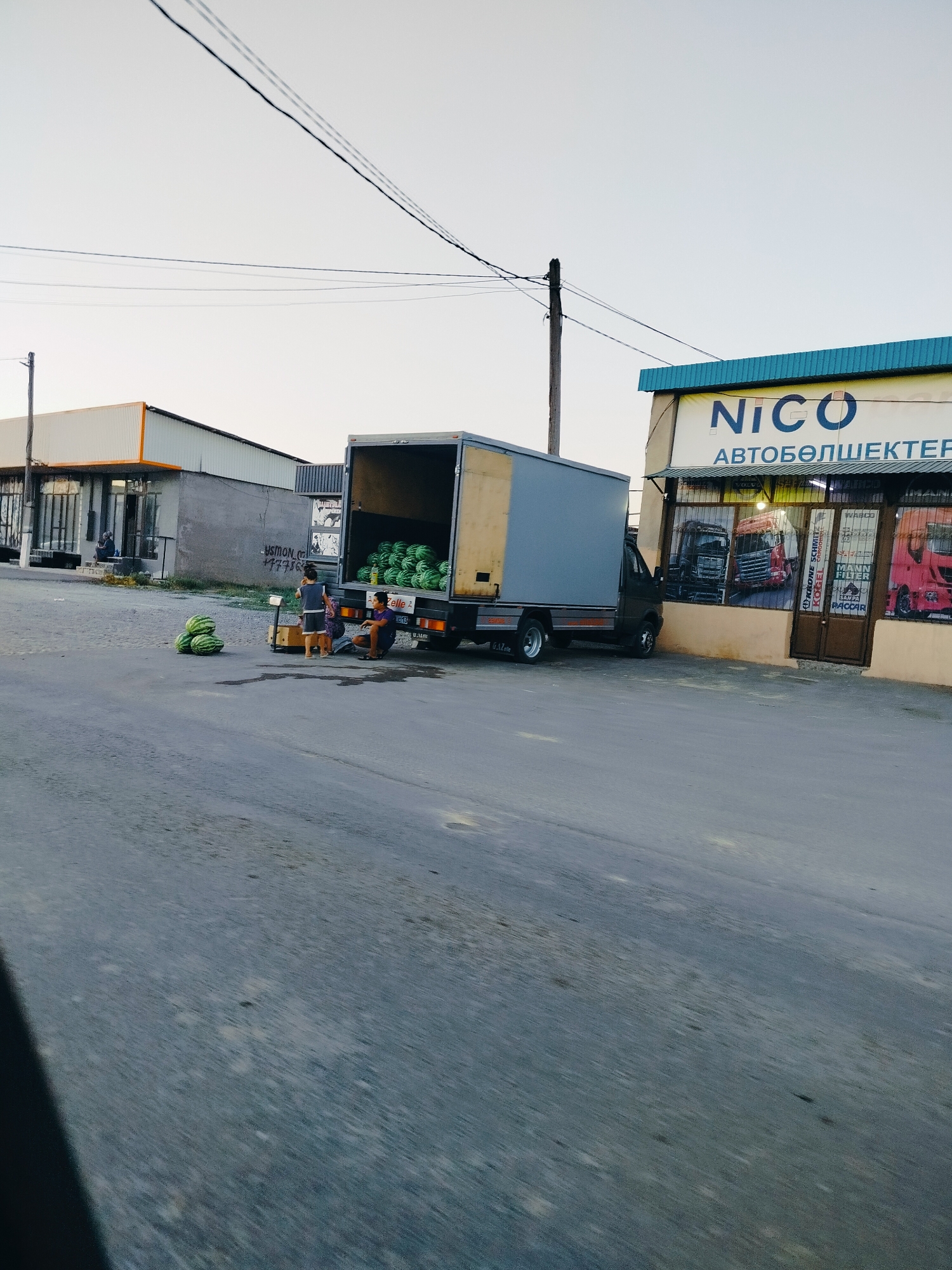 The image depicts a street scene in the city of Көлкент, Kazakhstan. In the foreground, the asphalt road is visible. To the left, several watermelons are piled on the ground. In the middle ground, a small group of people, including two adults and one child, are gathered around a scale. A truck with an open cargo area filled with watermelons is parked to the right of the people. Behind the truck, a building with a sign that reads "NICO АВТОБӨЛШЕКТЕР" is visible, suggesting it is an auto parts store. The sky is clear and appears to be late afternoon or early evening. The overall environment suggests a marketplace or a roadside stall where goods are being sold.