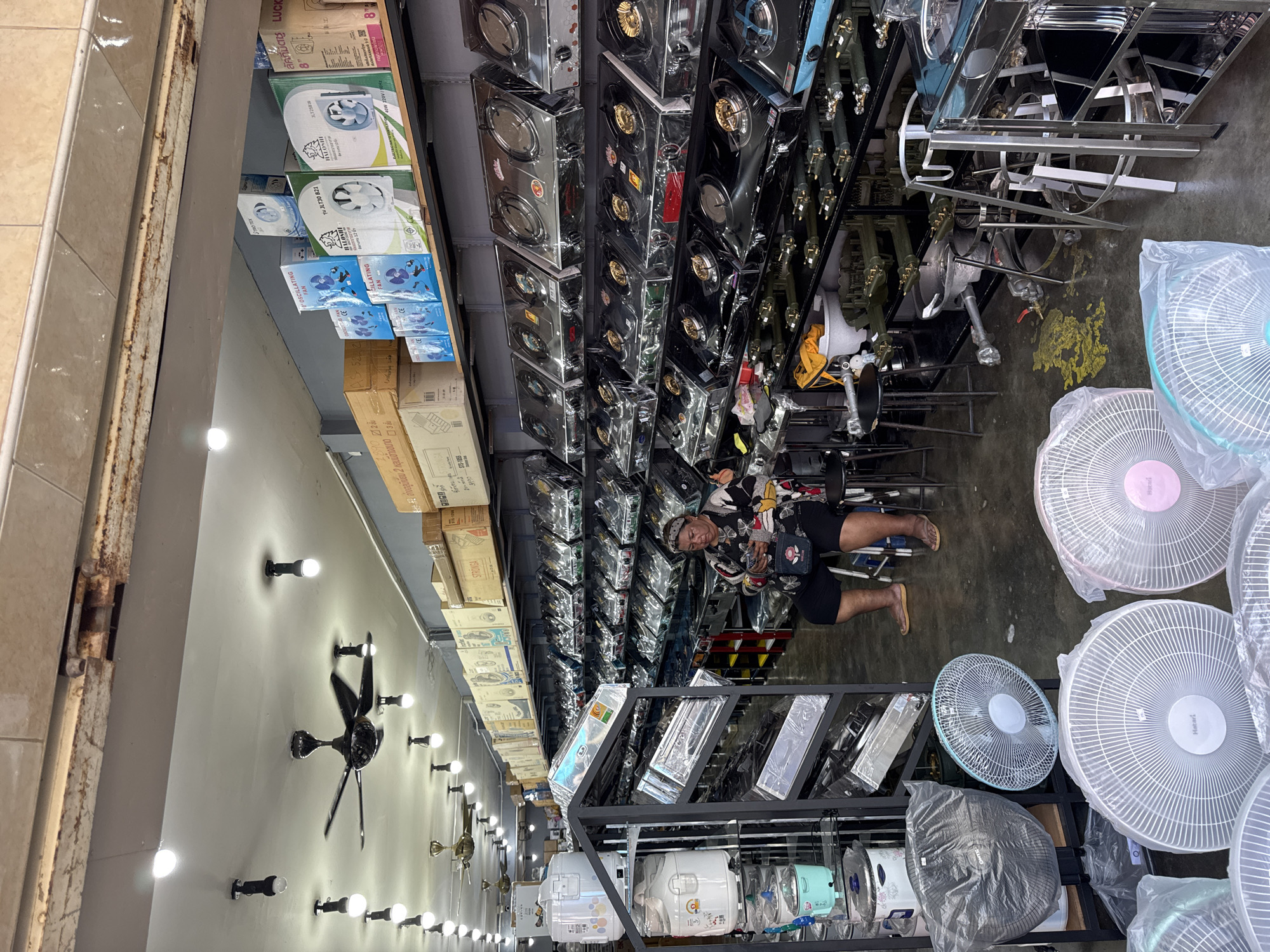 The image shows the interior of a retail store in Ranong, Thailand, specializing in kitchen appliances. The store is brightly lit with overhead fluorescent lights and ceiling fans. Rows of gas stoves are displayed on shelves, most covered in plastic wrapping. Various kitchen appliances, including rice cookers and electric fans, are also visible throughout the store. A woman is seated on a stool near the shelves of stoves, looking at her phone. The floor is concrete and appears to have some yellow staining. The overall environment suggests a busy marketplace or commercial district within the city.