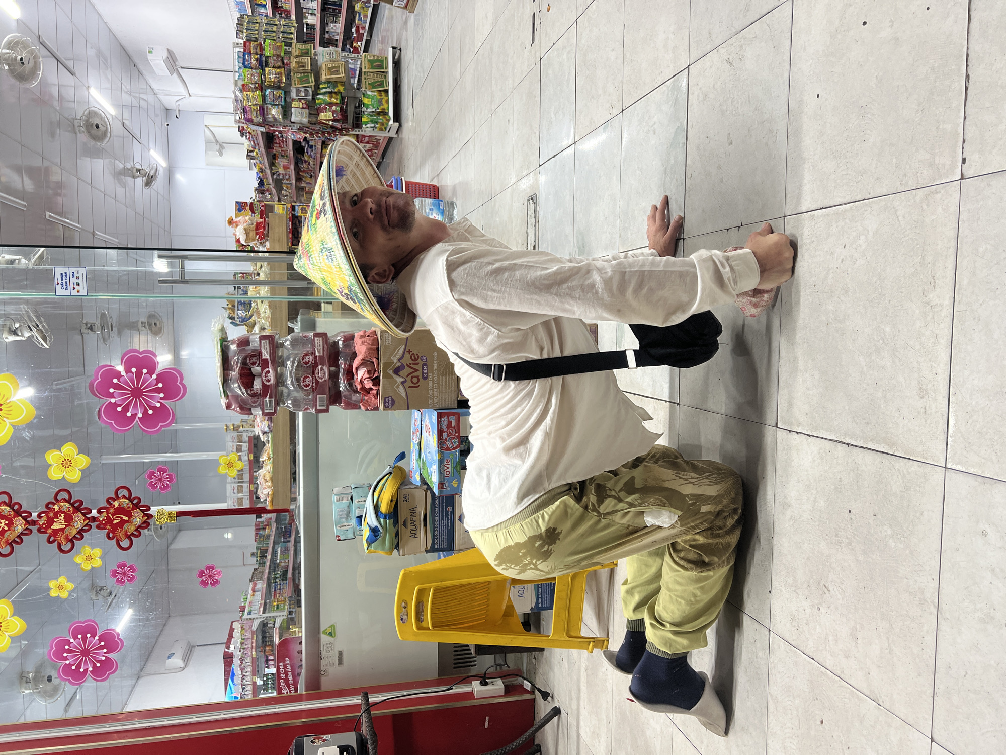 A person wearing a conical hat and a light-colored shirt is on their hands and knees on the tiled floor of a convenience store in Da Nang, Vietnam. The person is looking towards the camera with a slight smile. The store is well-lit with shelves stocked with various products. Decorations, possibly for a holiday, are visible on the glass storefront. A yellow plastic chair is positioned to the left of the person, and stacks of bottled water and other goods are visible in the background.