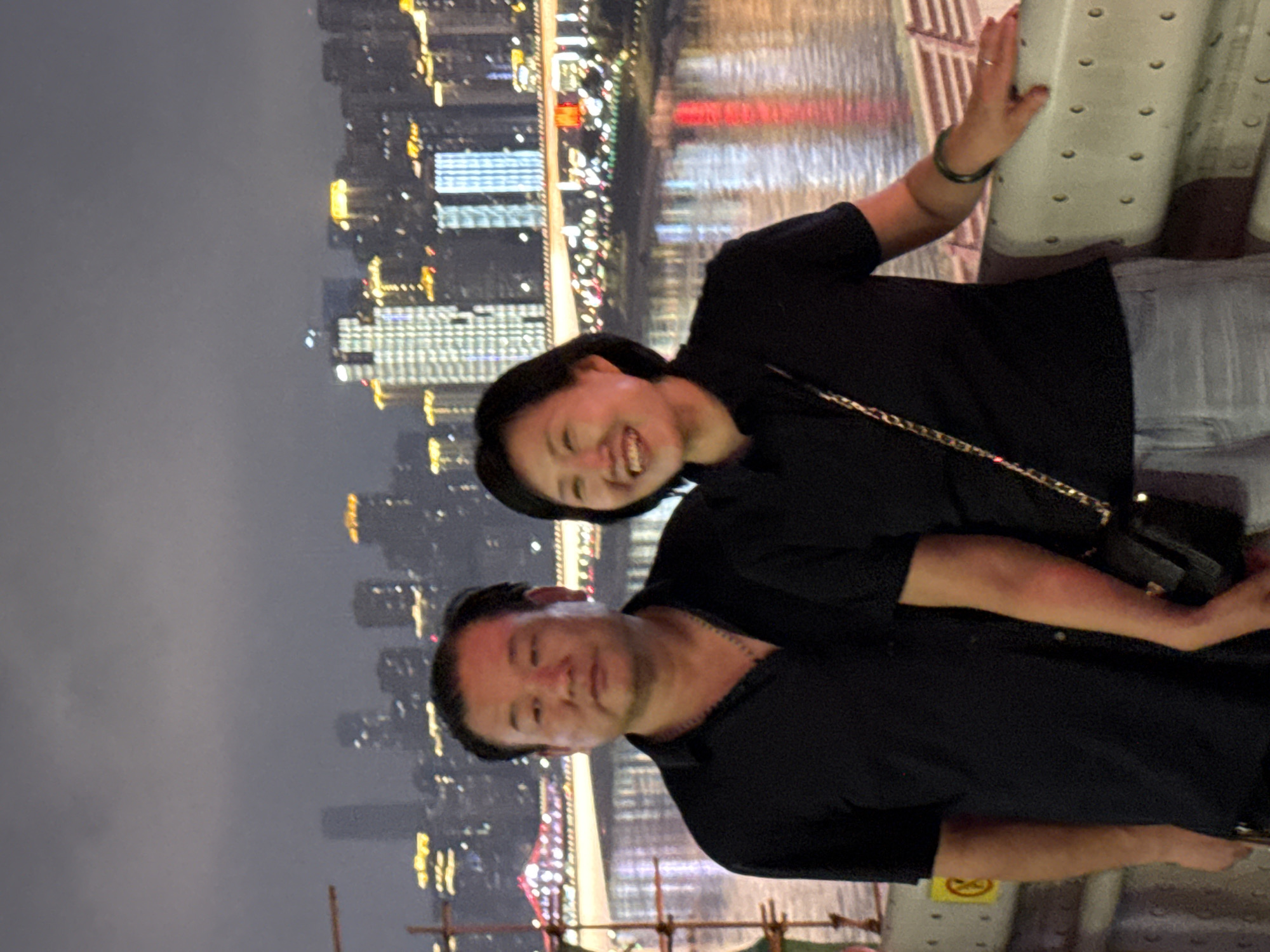 An Asian couple stands on a bridge with a city skyline in the background. The man, on the left, wears a black collared shirt and a necklace. He has dark hair and a round face. The woman, on the right, wears a black t-shirt and light-colored shorts. She has dark, short hair and is smiling. Her right hand rests on a white railing. A black purse with a gold chain is slung over her shoulder. The background shows a river reflecting city lights, buildings, and illuminated bridges. The scene is set at night, likely in Chongqing, Yuzhong District, China, given the distinctive urban landscape and bridge architecture.