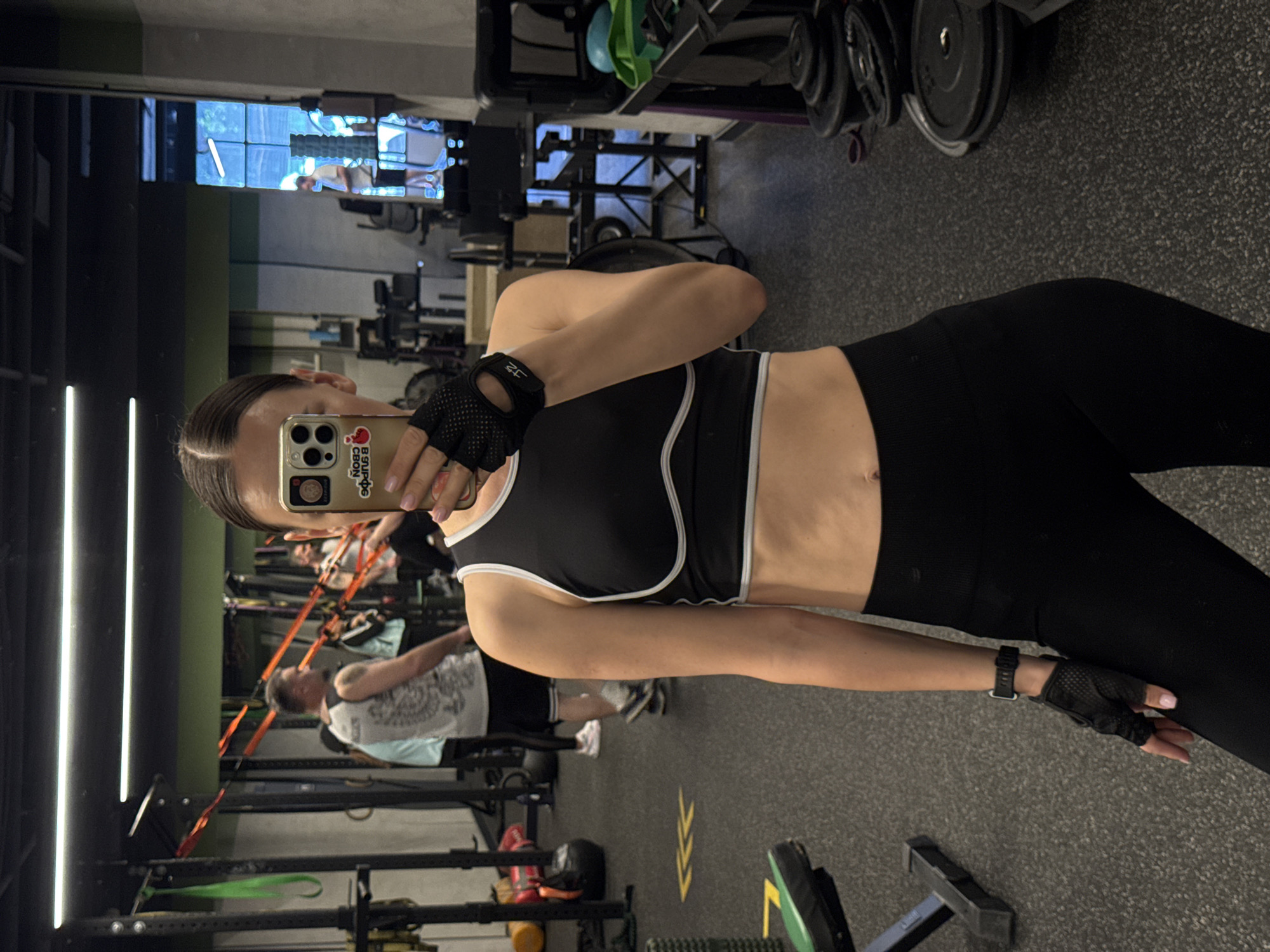 The image captures a female taking a mirror selfie in a gym located in Sochi, Russia. She stands slightly to the right, dressed in a black sports bra with white trim, black leggings, fingerless gym gloves, and a smartwatch on her left wrist. Her dark hair is pulled back, and her face is mostly obscured by the smartphone used for the selfie. The phone's light-colored case features a boxing glove with a heart icon and text that reads "Всигда свой Свои". The fingerless gloves have "J2" marked on them.
The reflection in the mirror reveals a bustling gym environment. In the background, another man with short hair, wearing a light-colored t-shirt, is visible exercising with orange resistance bands attached to a wall-mounted rack. Various gym equipment is scattered around, including stacked weight plates, a black weight bench, a green resistance band, and a roller. The gym features dark, textured flooring with yellow directional arrow markings. Vertical strip lights illuminate the left side of the mirror, and the walls are a mix of green and dark tones. The scene depicts an indoor fitness setting, likely during the day or under bright artificial lighting.