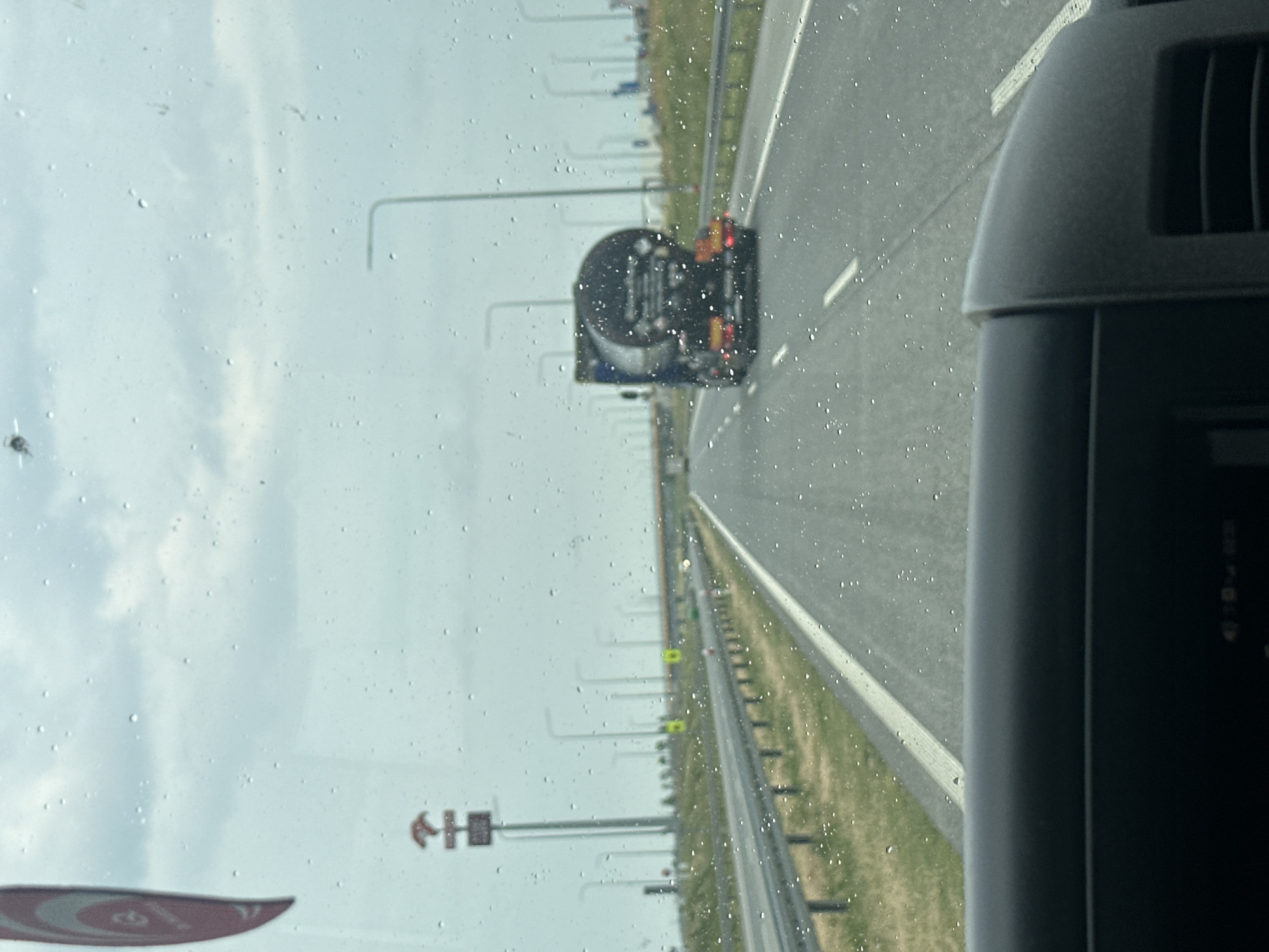 The image shows a view from inside a vehicle looking out through a rain-streaked windshield at a highway. A dark-colored tanker truck is traveling ahead on the right lane of the highway. The sky is overcast. On the left side of the road, there are streetlights and a signpost with a circular logo and a stylized “H”. The scene appears to be on a highway in or near Chomentowo, Poland, based on visual cues and context. The dashboard of the vehicle is visible in the foreground.
