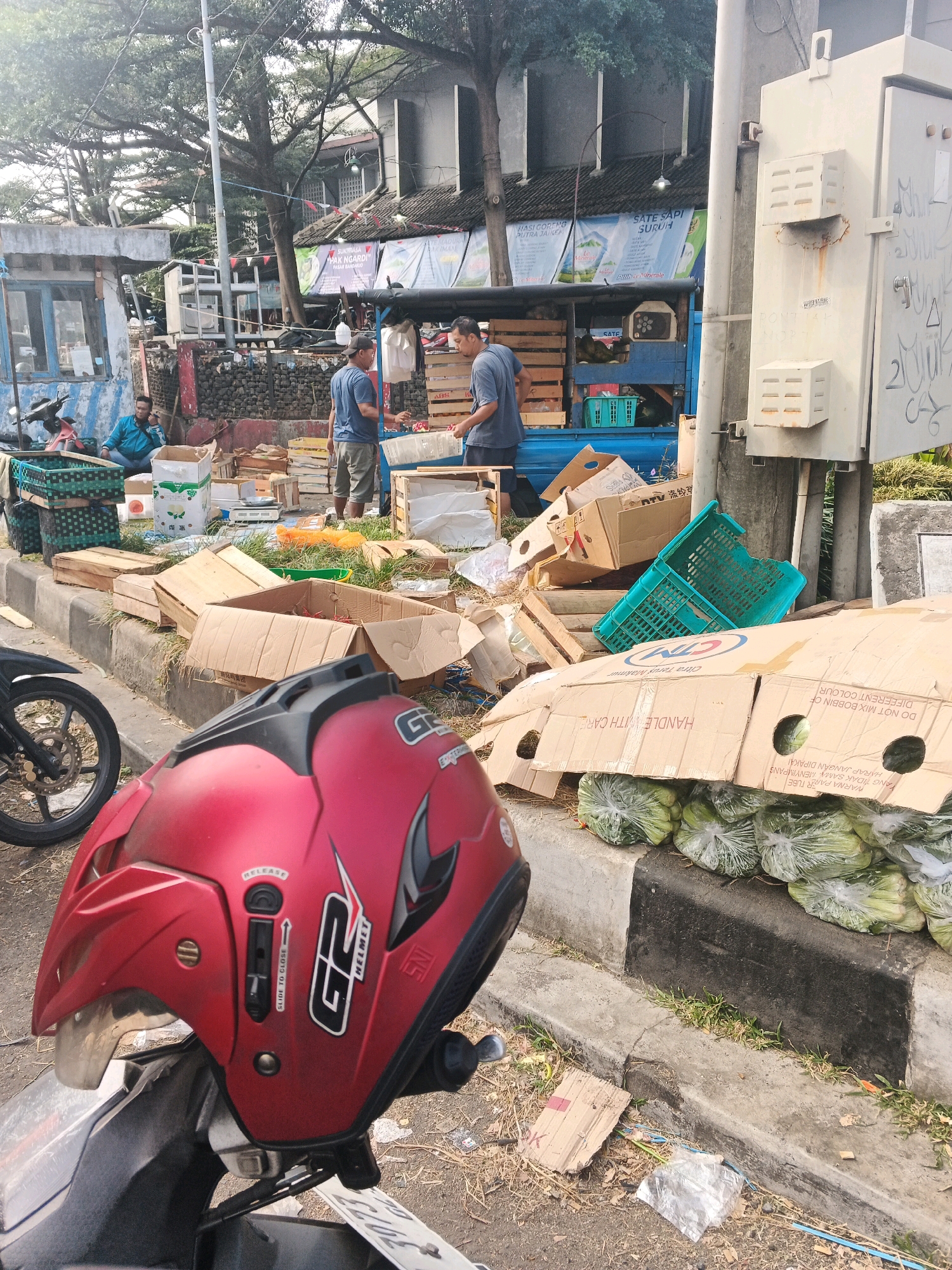 In Ungaran, Indonesia, an outdoor market scene unfolds. Two men stand near a bright blue truck, appearing to be engaged in activity related to produce. One man, wearing a blue t-shirt and a cap, is facing away from the camera. The other man, in a lighter blue t-shirt, is facing the camera, with a slight smile. Behind them, the truck is partially loaded with boxes and crates, suggesting commerce.
To the left, a man in a turquoise shirt sits on a stack of green crates, looking to his right, perhaps observing the scene or waiting. Further left, a red motorcycle is parked, its front wheel visible, and a red helmet rests on its handlebars, indicating recent or imminent use.
The ground is cluttered with cardboard boxes, some open and some stacked, along with loose produce and plastic bags. A significant portion of the foreground is dominated by the red helmet, its visor up, obscuring part of the motorcycle's body.
In the background, buildings with tiled roofs and trees with sprawling branches suggest an urban setting. A grey electrical box is prominently mounted on a pole to the right, covered in graffiti. The overall impression is of a busy, informal marketplace, likely a daily occurrence in this Indonesian city.
