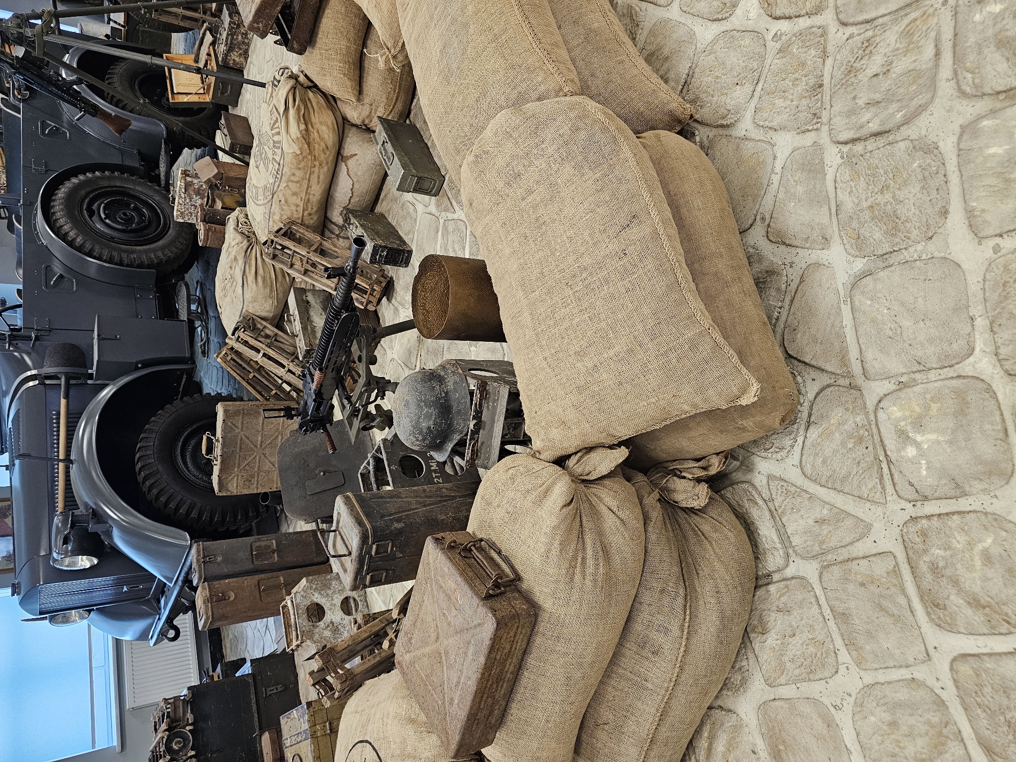 The image presents a collection of military artifacts and equipment arranged against a stone-tiled floor, possibly an exhibit or museum display. Dominating the scene are several burlap sacks, stacked and bundled together. Adjacent to these, there's an array of military-related objects including ammunition crates, metal containers (possibly fuel canisters), a soldier's helmet, and what appears to be a machine gun mounted on a stand. A vintage military vehicle, possibly a truck, is partially visible on the left side of the image, showcasing its tires and front section. The setting is likely an indoor environment. This image shows a display at the Vadim Zadorozhny Technology Museum in Архангельское, Russia.