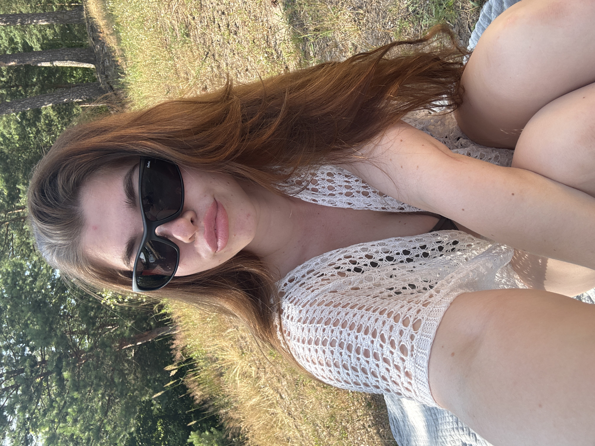 A young woman with long, reddish-brown hair and light skin is sitting on a blanket outdoors. She is wearing black sunglasses and a white crocheted top over a dark bikini. The background shows a grassy slope with trees, suggesting a park or natural area. The lighting indicates it is daytime, likely sunny. The location is identified as Kurovskoye, Russia.