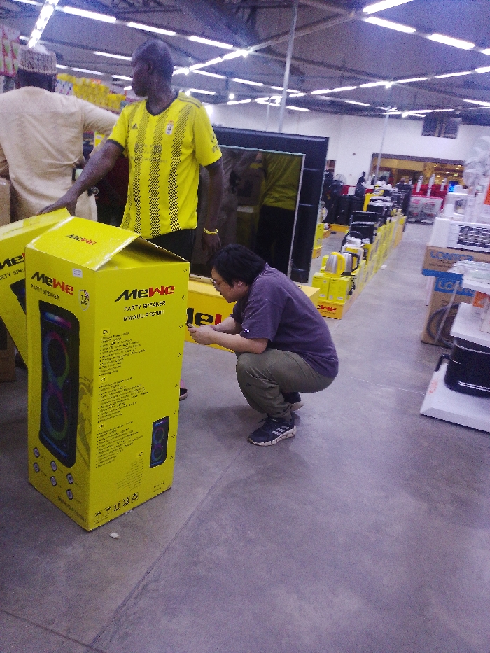 The image captures an indoor scene within an electronics retail store in Gandu Albasa, Nigeria. Three individuals are prominently featured. In the foreground, a person of East Asian descent, wearing a dark purple short-sleeved shirt, olive green cargo pants, and black and white athletic shoes, is in a full squat position, focused on a smartphone held in both hands. To their left, an adult male of African descent stands upright, dressed in a yellow and black striped short-sleeved athletic shirt with a white emblem and a yellow wristband. He gazes towards the right of the frame, with his left hand resting near two large, bright yellow cardboard boxes. These boxes are labeled "MeWe PARTY SPEAKER MWAUD-PTS100"; one is partially open, revealing a dark speaker unit with multi-colored lights. In the background, partially obscured, an adult male of African descent wearing a light beige traditional Nigerian garment and cap is seen facing away, appearing to interact with merchandise on a display. The store environment includes a plain concrete floor, a ceiling with visible ductwork and evenly spaced fluorescent strip lighting, and multiple rows of electronics, including large flat-screen televisions and smaller appliances displayed on yellow stands and shelves, extending into the background.