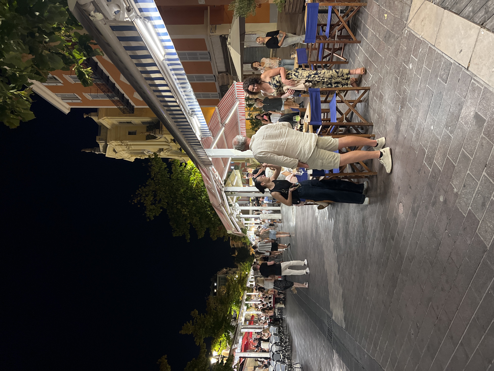 This is a nighttime exterior shot of the Cours Saleya market in Nice, France, from a slightly elevated perspective. The cobbled pedestrian walkway is lined with outdoor seating for restaurants. In the foreground, two men and two women are standing near tables with blue chairs. The man on the left is wearing a light-colored short-sleeved shirt and shorts, while the woman next to him is in a black top and dark pants. To their right, a man in a light tan shirt and shorts stands facing away from the camera, and a woman in a patterned dress is facing him. Behind them, other diners are seated at tables under striped awnings. The buildings lining the walkway are illuminated, with orange and yellow facades visible. Trees with green foliage are scattered throughout the scene, and the night sky is dark.