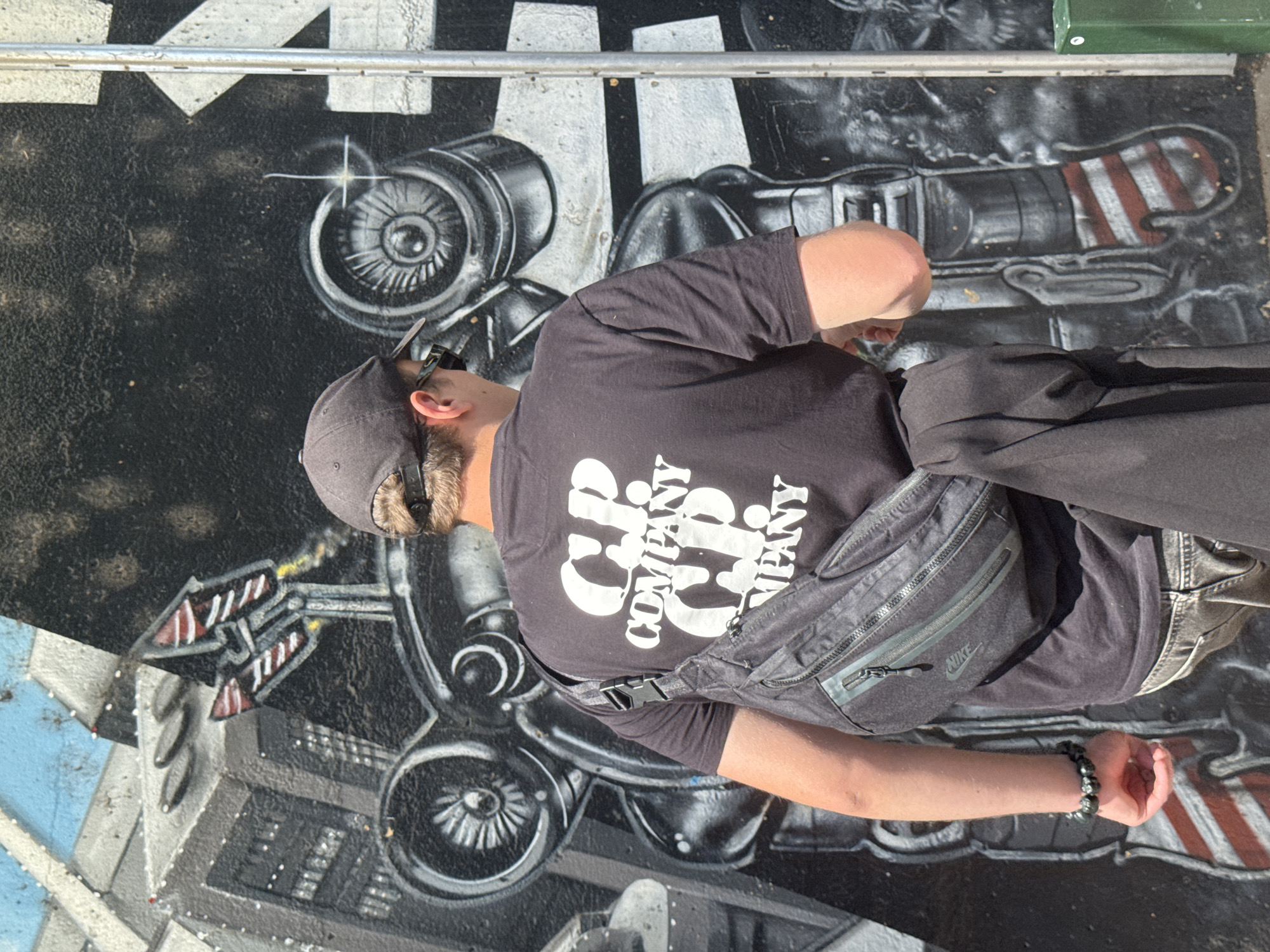 A young person, from the rear, stands in front of a graffiti-covered wall, in an outdoor urban setting. The graffiti depicts a large, mechanical robot-like figure and city structures. The person wears a dark grey baseball cap, sunglasses, a black t-shirt with white "C.P. COMPANY" branding repeated on the back, dark grey jeans, and a dark grey sling bag worn across their chest. The person has a bracelet on their left wrist. A dark jacket is draped over their right arm. The image appears to be taken on a sunny day. The graffiti style suggests this location could be in the city of Brussels, Belgium, which is known for its vibrant street art scene.