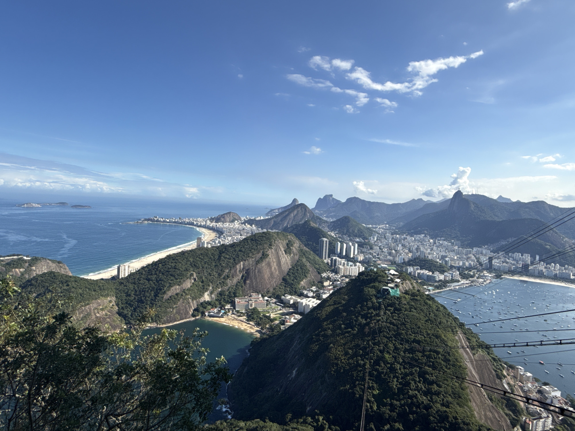 This panoramic high-angle photograph captures a breathtaking view of a vibrant coastal city, likely Rio de Janeiro, nestled between towering green mountains and the expansive blue ocean under a clear sky.
In the foreground, the steep, densely vegetated slope of a mountain dominates the right side, with several parallel cable car lines stretching diagonally across the frame, hinting at an aerial tramway system. Below, a small bay with a sandy beach and some buildings is visible.
The midground unfolds a dramatic landscape. To the left, a long, curving white sand beach, lined with numerous high-rise buildings, meets the deep blue sea. The ocean extends to the horizon, dotted with a few small islands. In the center, another prominent, rocky, tree-covered mountain rises, with urban development clustered around its base. To the right, a large, sheltered bay (likely Botafogo Bay) is filled with hundreds of small boats and yachts anchored in its calm waters, bordered by a bustling cityscape.
The background features a majestic series of rugged, verdant mountains characteristic of the region, towering over the urban sprawl that continues to extend into the distance along the coast and valleys. The sky above is a brilliant azure, adorned with scattered white cumulus clouds, indicating a bright and clear day.
The scene depicts a bustling urban environment harmoniously integrated with stunning natural beauty. No specific human activity is discernible beyond the general presence of a city, boats, and the cable car infrastructure. There is no visible text in the image.
