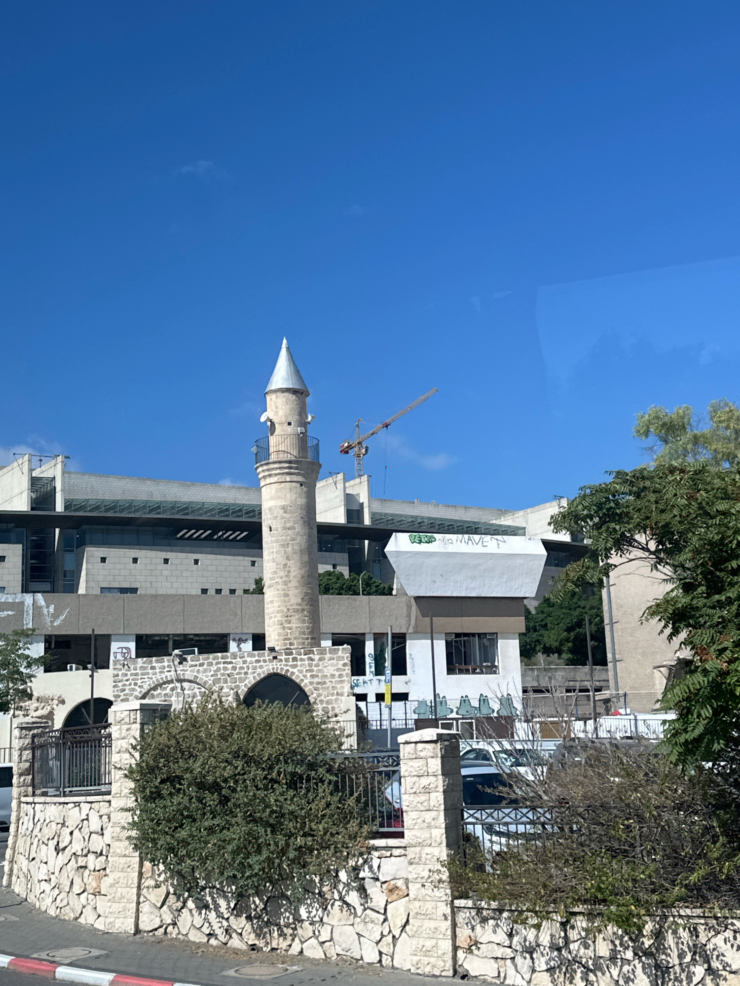 The image shows a minaret of a mosque in the city of Haifa, Israel. The minaret is made of light-colored stone and has a conical roof. It is surrounded by modern buildings, including a large concrete structure with glass windows. A construction crane is visible in the background. The sky is clear and blue, and there are some trees and bushes in the foreground. The minaret appears to be part of a complex that includes an arched entrance and a stone wall. There is graffiti on some of the buildings.