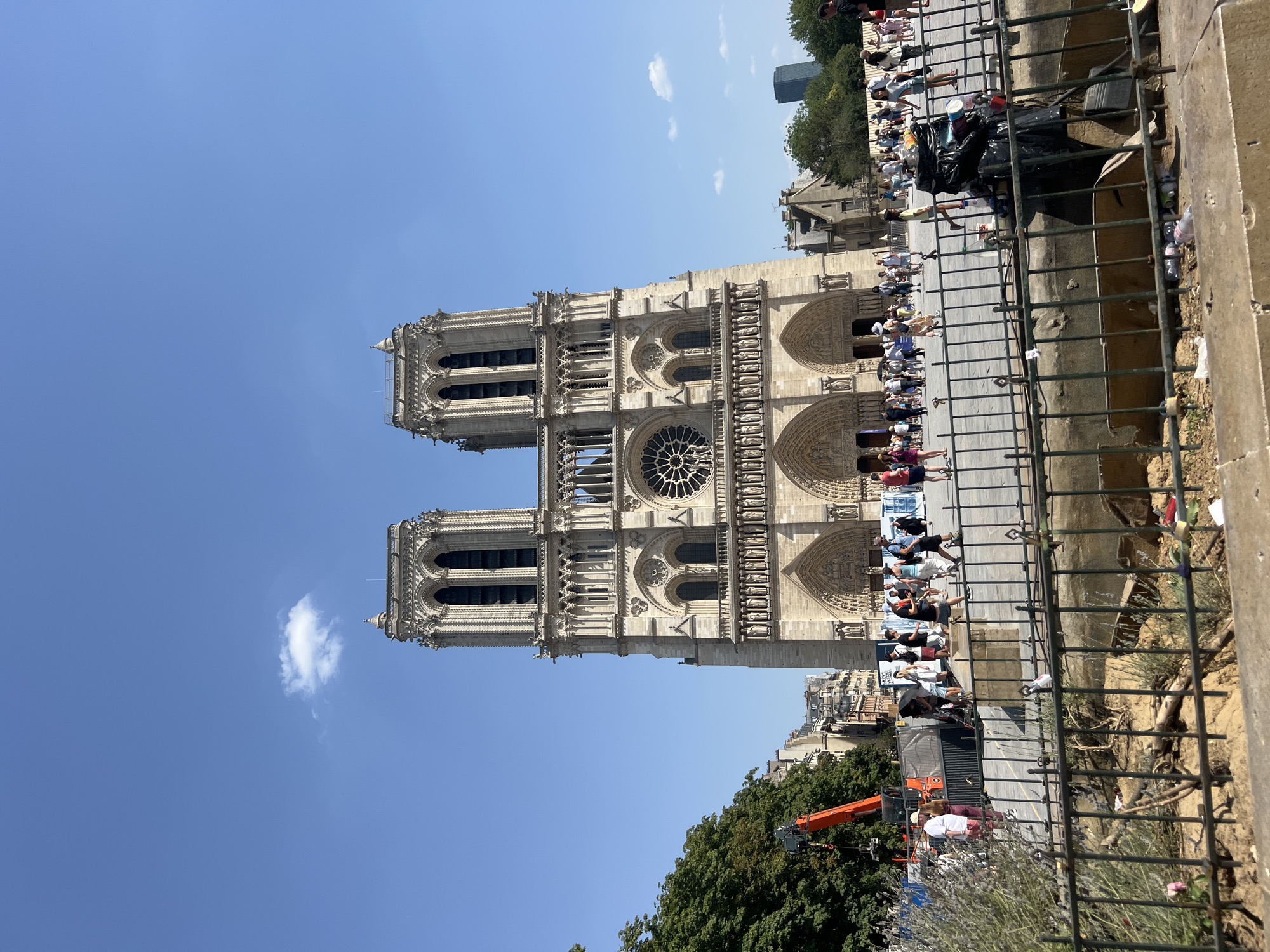 The media file captures a view of Notre Dame Cathedral in Paris, France, under a clear blue sky with a few scattered clouds. The cathedral's facade, featuring intricate architectural details like arched doorways, statues, and a large rose window, is prominently displayed. Two towers rise symmetrically from the front of the building.
In the foreground, a metal fence separates the viewer from a plaza area. Beyond the fence, a crowd of people is gathered, appearing to be tourists or onlookers observing the cathedral. Some individuals are standing, while others are seated or walking. Their attire suggests a warm, sunny day, likely in the spring or summer.
Behind the crowd and to the right of the cathedral, trees and other Parisian buildings are visible, providing context to the urban setting. There is also construction equipment, including an orange crane, suggesting ongoing work or restoration efforts at or near the cathedral. The presence of trash bags and some scattered litter near the fence indicates that this is a public space with regular foot traffic.
The overall impression is a daytime scene, likely late morning or early afternoon given the bright sunlight and clear skies. The mood appears to be one of appreciation for the historical landmark, with a blend of sightseeing and the routine activities of urban life, including construction and public gathering. No discernible text is visible in the media file.