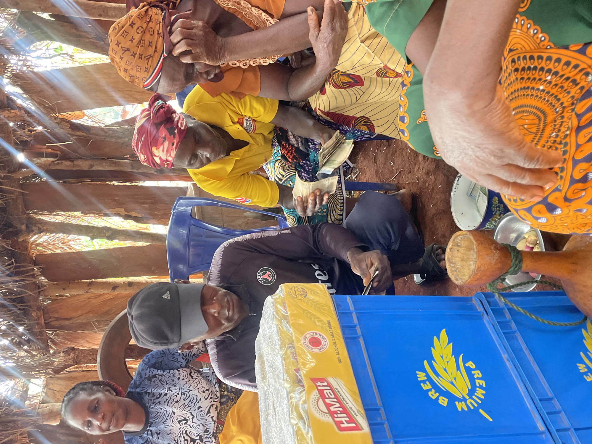 A group of people are gathered indoors in what appears to be a hut-like structure in Ogbe, Nigeria. Four women are visible wearing colorful traditional clothing and head coverings. One woman is handling what appears to be money. A man in a black jacket and cap sits among them, looking at something in his hands. In front of him are crates labeled "Premium Brew" and "Hi-Malt," suggesting a beverage distribution or sales point. The interior walls are made of wooden planks. Sunlight streams through the spaces between the planks. There are metal plates. A gourd is also visible.