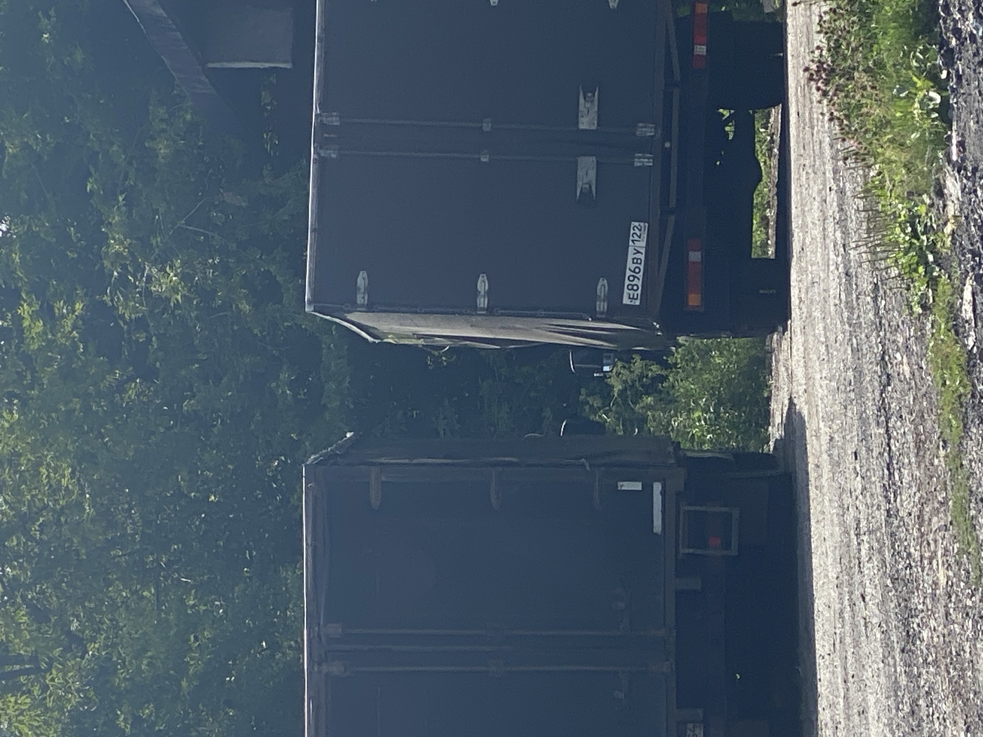Two dark-colored trucks are parked on a gravel surface, with dense green foliage in the background. The truck on the right has a license plate reading "E896BY 122". The scene appears to be in an outdoor setting, possibly a rural area or a less developed part of a city. The lighting suggests it is daytime.