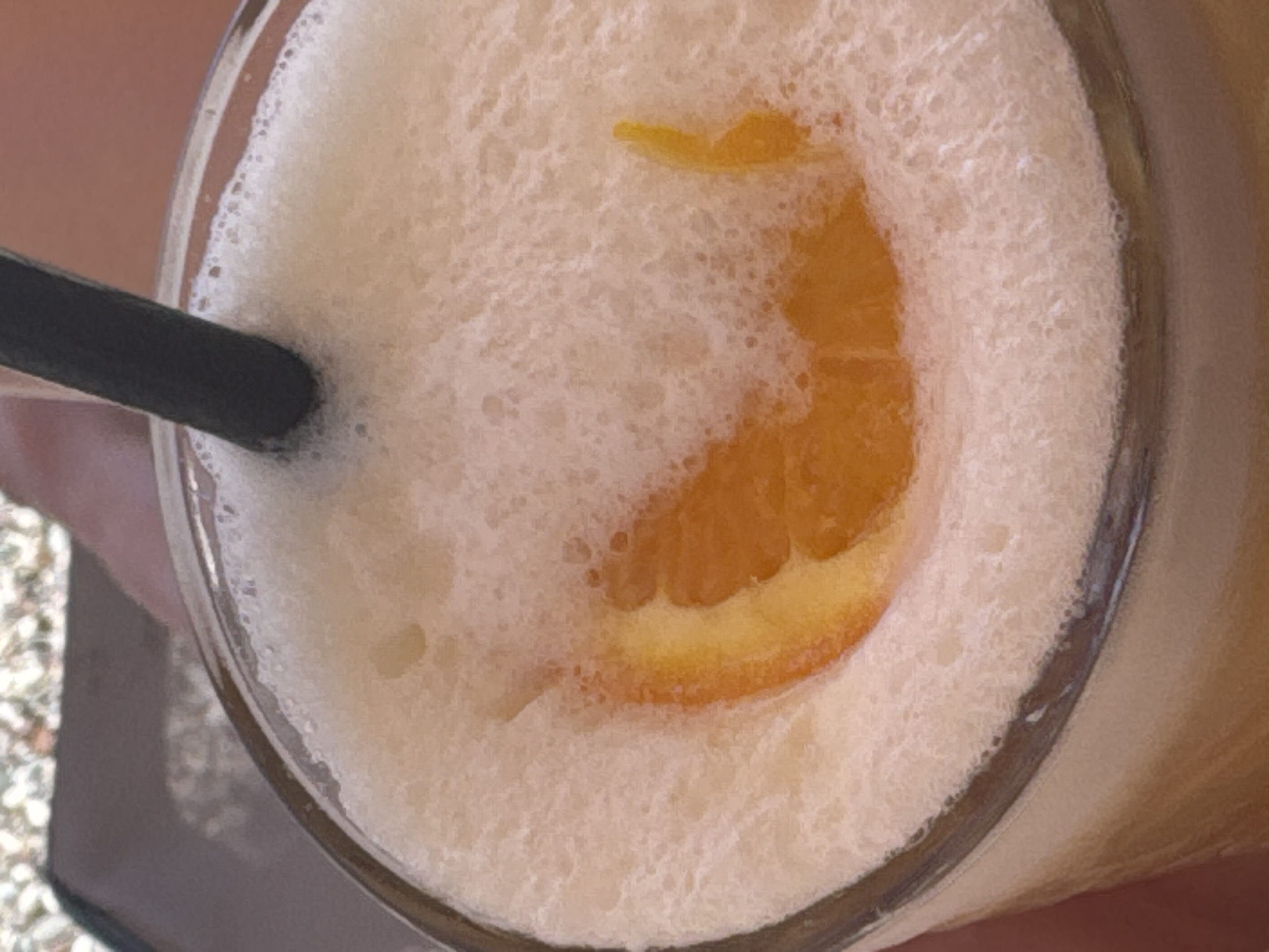 A close-up, overhead shot shows a glass filled with a foamy, light orange beverage. A dark straw is partially submerged in the foam, angled from the top right to the center of the frame. A thin slice of orange is visible submerged in the foam, appearing as a crescent shape with its bright orange pulp peeking through. The glass has a clear rim, and the background is out of focus, showing a textured surface that appears to be an outdoor table with pebbles. The overall impression is one of a refreshing drink, likely enjoyed outdoors. The location is Unknown, Unknown.