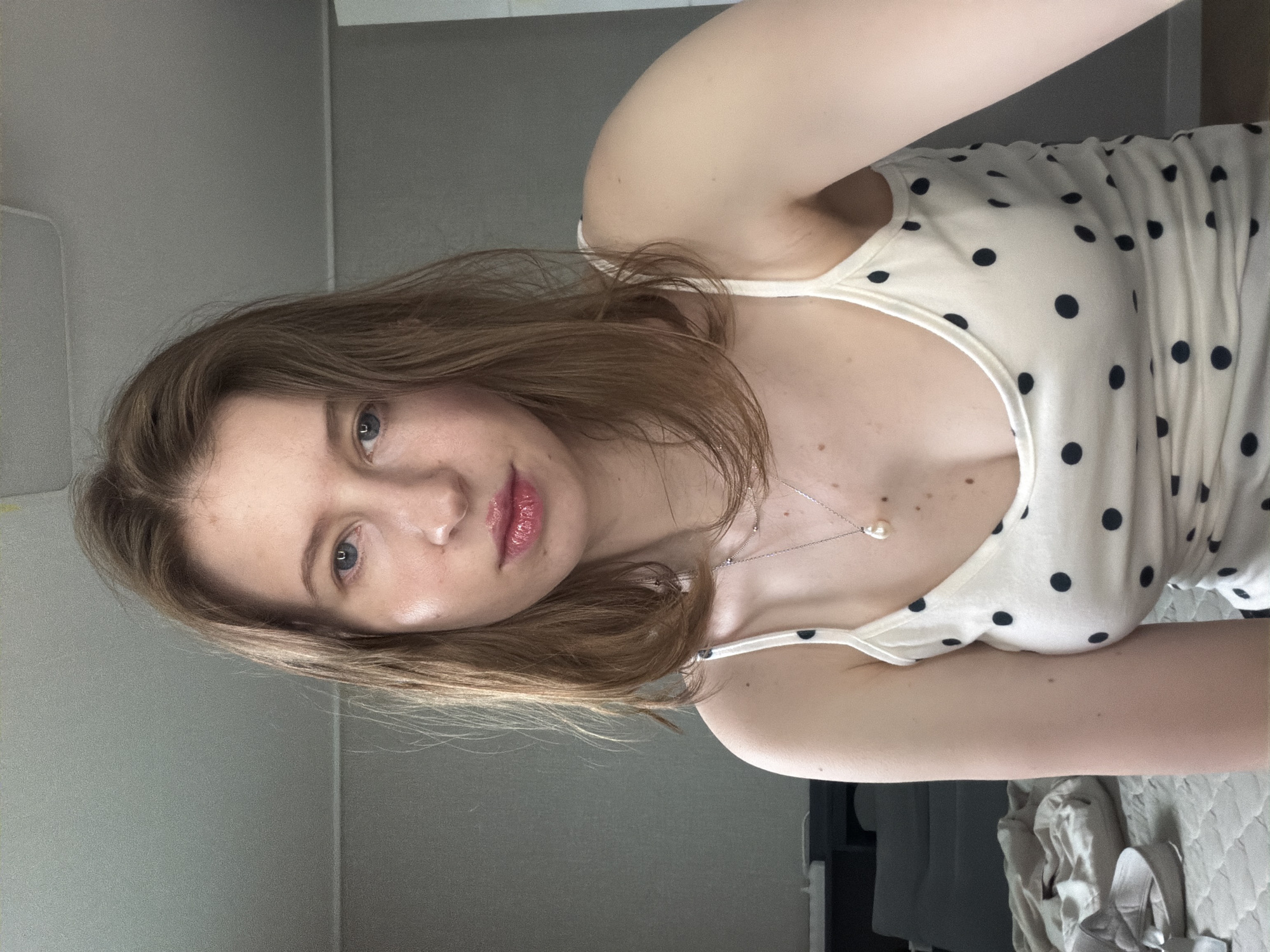 A woman is captured in a selfie, her head tilted to her left. She has shoulder-length, light brown hair and blue eyes. Her lips are glossy and pink. She is wearing a white tank top adorned with black polka dots and a delicate silver necklace with a single pearl pendant. The background appears to be an indoor setting, with a muted gray wall and a glimpse of a patterned surface, possibly a bed, in the lower right corner. The lighting suggests it could be daytime, given the natural light illuminating her face. Her expression is neutral, with a slight smile. The image appears to be taken in Dongducheon-si, South Korea.