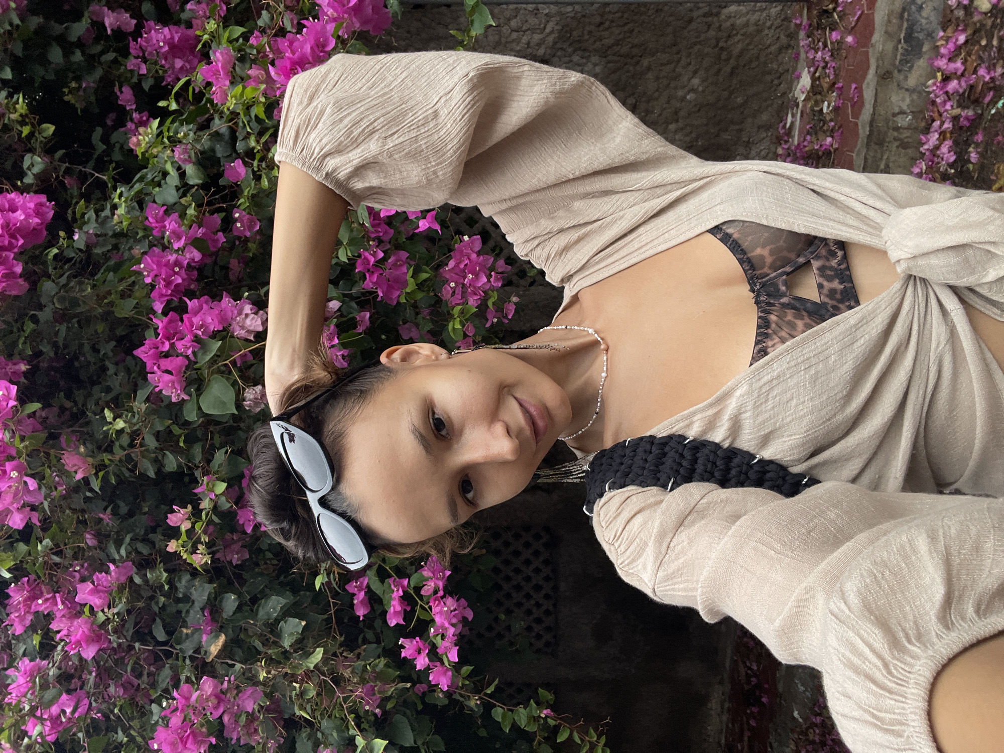 A woman with sunglasses atop her head poses for a photo next to a bougainvillea bush in Phu Thuy Ward, Vietnam. She has fair skin, brown hair, and wears a silver necklace. She is wearing a loose-fitting tan shirt that is knotted at the waist, revealing a patterned bra. A thick black knitted strap crosses her shoulder. The bougainvillea bush is dense with green leaves and bright magenta flowers. A rough concrete wall is visible in the upper right corner.