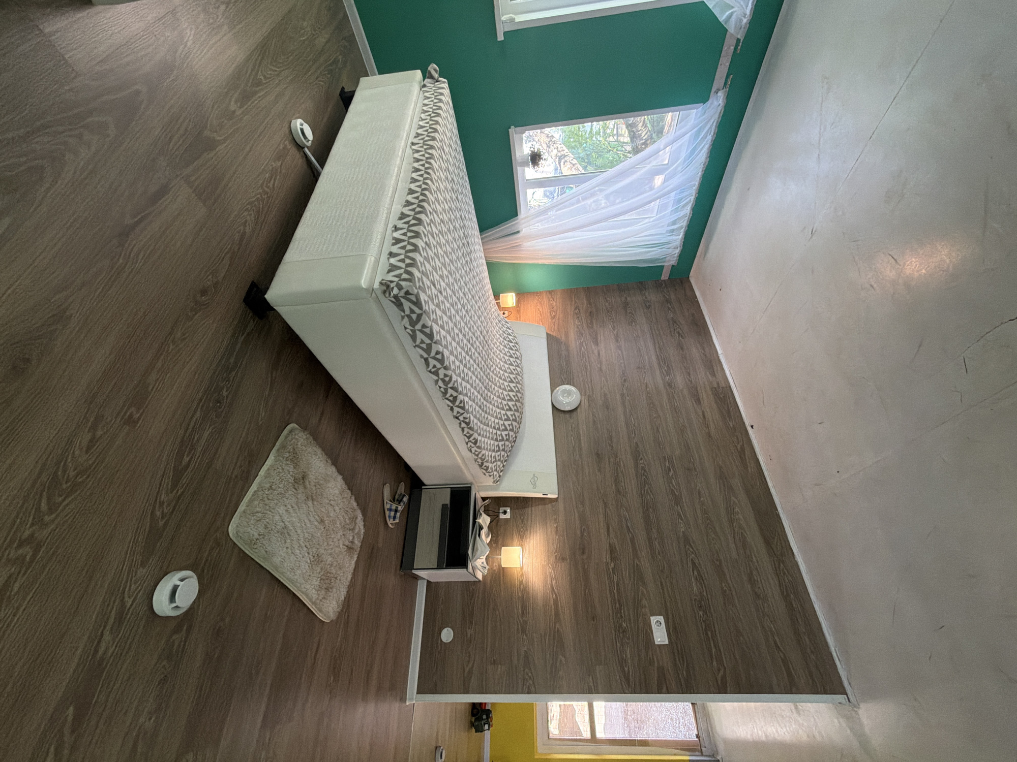 The image depicts an interior scene, likely a room, that is inverted, giving the impression that the ceiling is the floor and vice versa. A large, white bed with a patterned comforter is suspended from what would normally be the ceiling. To the left of the bed, attached to the "ceiling," is a dark cabinet with a mirrored front. Below this, on the "wall," there are two light fixtures and a smoke detector. A small, fluffy rug is also affixed to the "ceiling." The "walls" appear to be paneled with wood-grain material and painted a vibrant green. A window with sheer white curtains is visible on the green wall. The "floor" is a light-colored, smooth surface. A doorway leading to a yellow-painted area is partially visible on the left. The overall impression is of a deliberately disorienting space, characteristic of an "upside-down house" attraction. This specific location is not identifiable from the image alone and cannot be confirmed to be in Izhevsk, Russia.