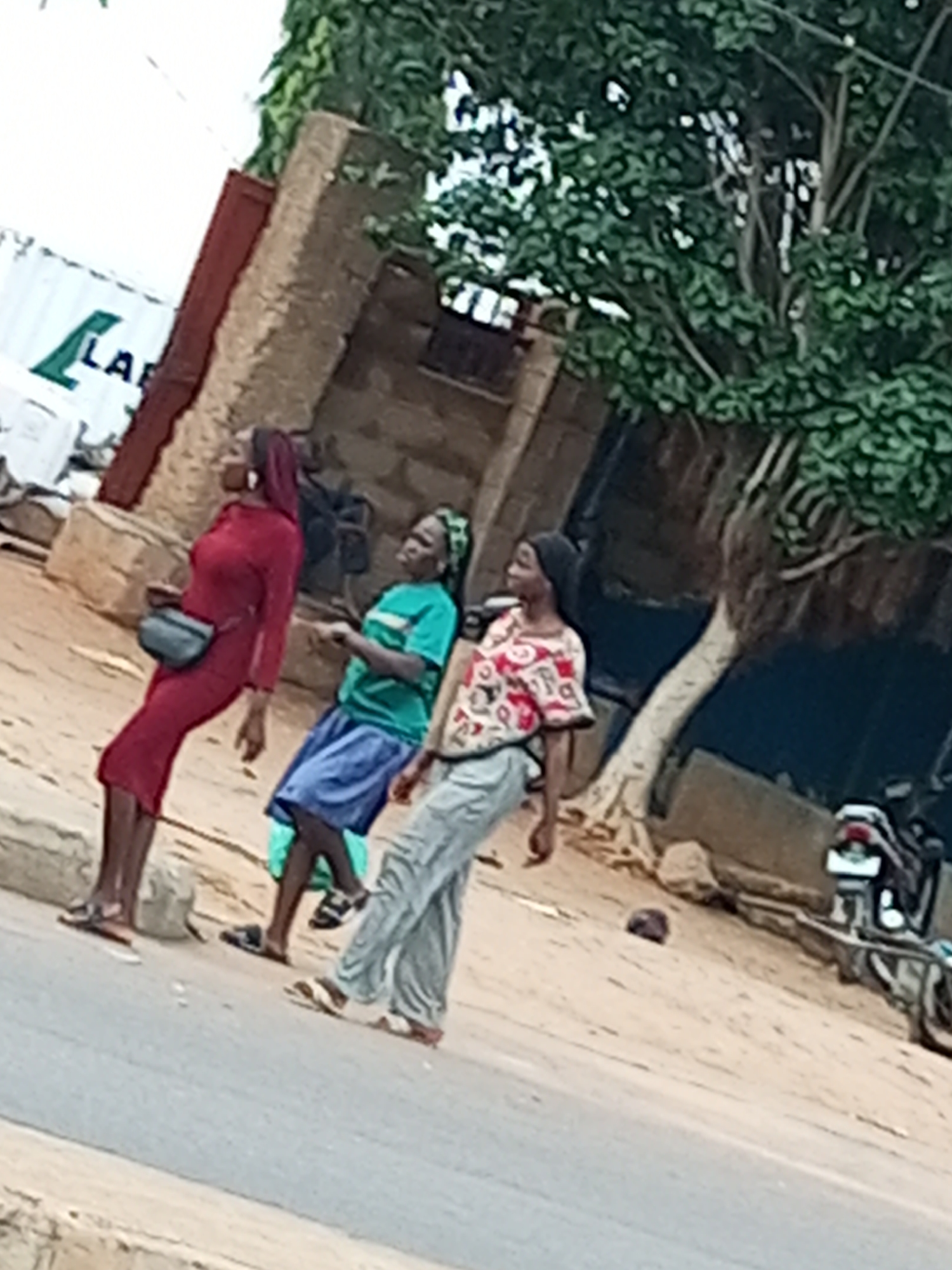 Three people are walking along a street in Tumfure, Nigeria. The person on the left is a woman wearing a long, form-fitting red dress and a dark fanny pack. She has dark hair, partially covered by a maroon headscarf. The middle person is wearing a green t-shirt and a blue skirt, with a green headscarf. The person on the right is wearing a patterned shirt with red, white, and black designs, and light grey pants. They are all wearing sandals.

The scene appears to be outdoors during the daytime, possibly late morning or early afternoon, with a tree providing some shade. The ground is a mixture of paved road and dirt. In the background, there is a brick building and a large white shipping container with green lettering that appears to read "LA." A motorcycle is parked on the right side of the image. The overall impression is a casual, everyday scene of people going about their business on a street. There are no strong indicators of specific emotions or a particular event taking place.