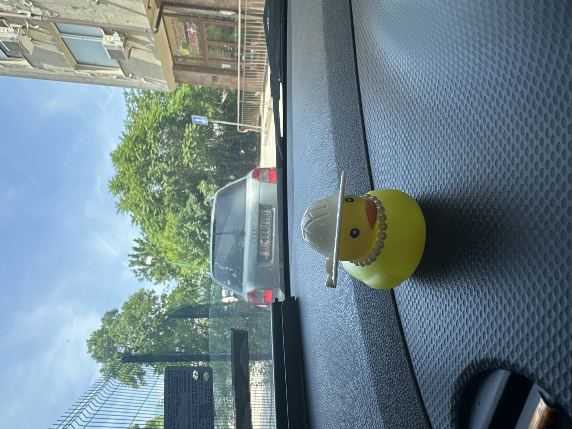 The image captures a static scene from inside a vehicle, looking through the windshield towards an outdoor urban environment under a clear blue sky. In the foreground, positioned centrally on a textured black dashboard, is a yellow rubber duck figurine. This duck wears a white, wide-brimmed hat and a white pearl-like necklace, featuring an orange beak and black eyes.
Through the windshield, a street scene unfolds. Directly ahead, a silver Audi sedan is parked, displaying the Bulgarian license plate "B 4440 HB" on its rear. To the right of the Audi, a multi-story building with a light-colored, weathered facade is visible, featuring several rectangular windows and external air conditioning units. The ground floor of this building houses a shop with a brown entrance and a sign displaying red and yellow text, partially legible as "КЕЙК+С". A metal railing stands in front of the shop.
On the left side of the view, a dark metal fence extends upwards, behind which dense green trees are visible. Additional trees are scattered across the background, beneath the clear blue sky. A blue square road sign with a white upward-pointing arrow is visible by the roadside, to the right. A sticker with Bulgarian text is partially visible on the upper left interior of the windshield. The scene depicts a daytime setting in the city of Unknown, Unknown.