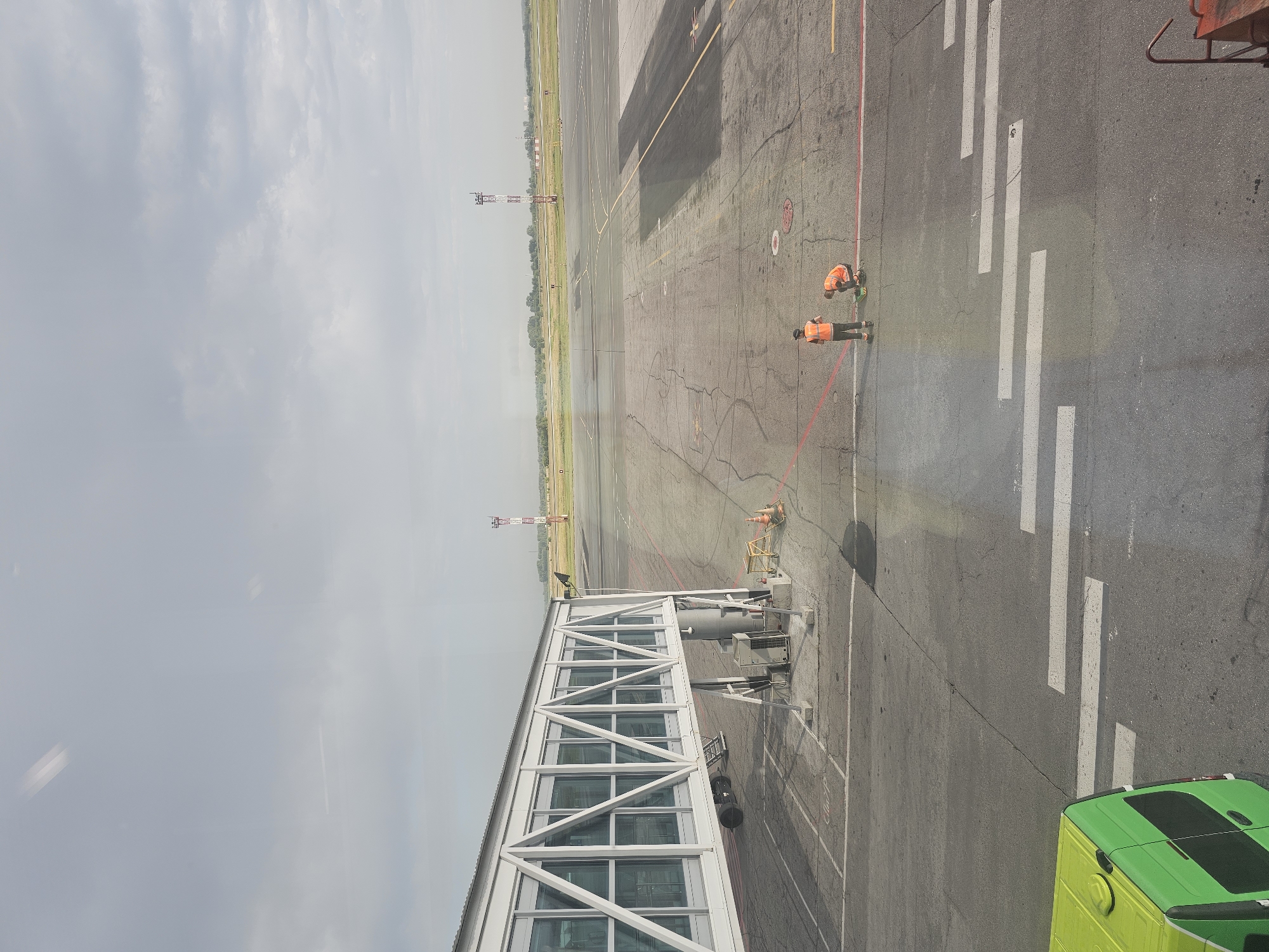 The image depicts an airport tarmac in the city of Ob, Russia. A modern, white jet bridge extends from a building on the left side of the frame towards the tarmac. The tarmac itself is paved and shows signs of wear, with various markings and some cracks. In the middle ground, two individuals wearing high-visibility orange vests are present on the tarmac, one standing and the other kneeling. They appear to be working on the ground, though their specific task is unclear. In the background, a tall, striped tower, possibly an air traffic control tower, is visible along with other airport infrastructure and grassy fields. A bright green vehicle is partially visible in the lower left corner. The sky is overcast with diffused light.