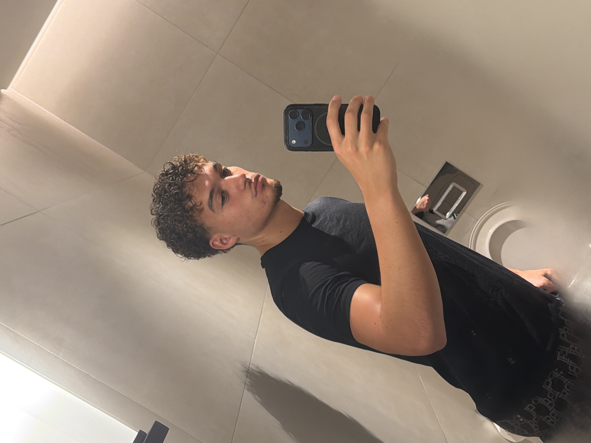 A light-skinned man with curly hair is taking a selfie in a bathroom. He is wearing a black t-shirt and holding a black smartphone in his right hand. A sink with a faucet and a toilet are visible in the background. The bathroom walls and ceiling are tiled in a light color. The location is not discernible as being in Dubai.