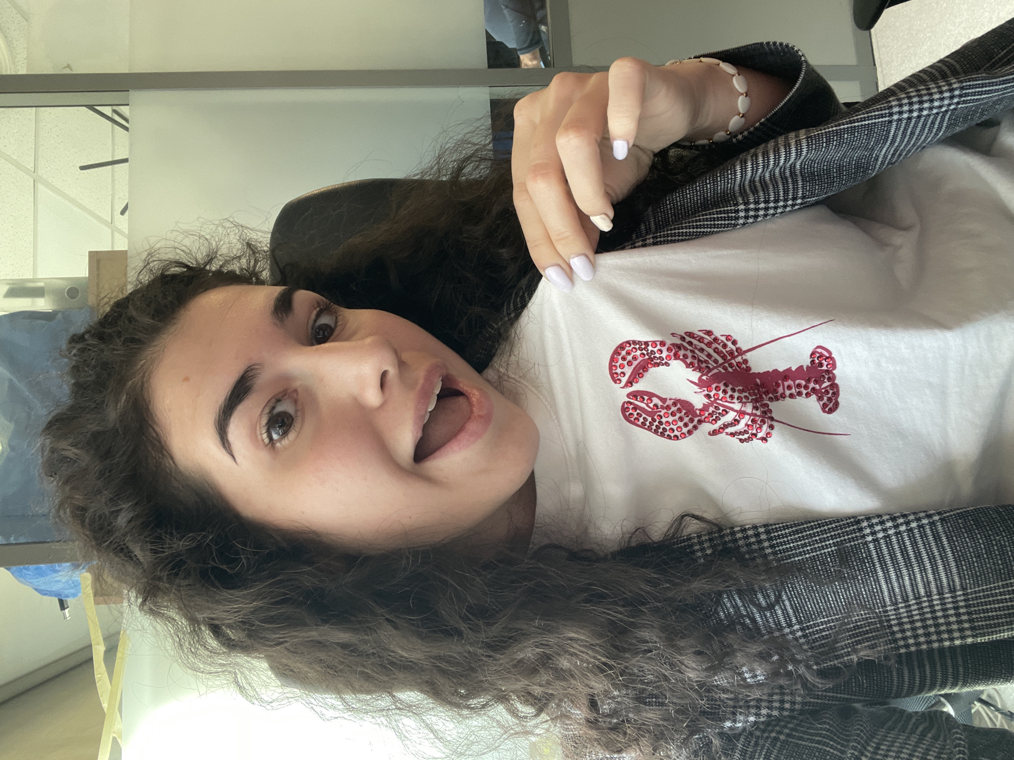 A woman with dark curly hair, wearing a white t-shirt with a red lobster design and a grey plaid blazer, is shown in a selfie. She has light skin and is smiling. Her nails are painted a light color. She is indoors, in a room with office-style windows and fixtures. The location cannot be determined from this image.