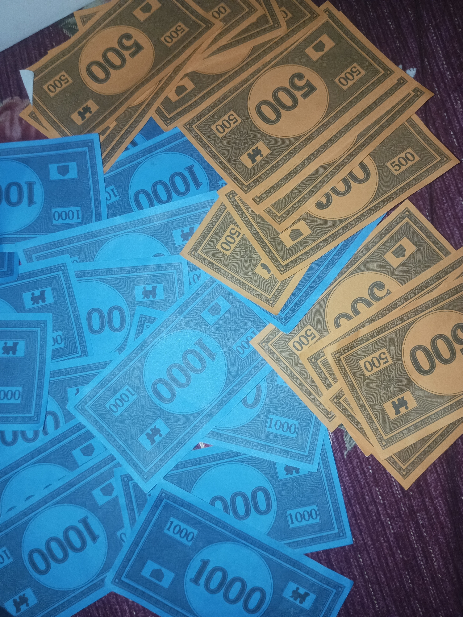 The image displays a scattered arrangement of board game money, specifically Monopoly currency, spread across a textured fabric surface, which appears to be a dark purple or maroon bedspread or blanket, likely within an indoor setting in Gwarinpa, Nigeria.
The money is differentiated by color and denomination. Blue bills are predominantly visible, featuring a large '1000' in the center, with smaller '1000' or '100' visible in the corners of some notes. Some blue notes also show a large '00' in the center, possibly indicating a '100' denomination. The tan/gold bills mostly display '500' as their central denomination, with smaller '500' or '50' visible in the corners of some notes.
Both the blue and tan/gold bills share a similar design, including a decorative border pattern and a small, characteristic symbol, resembling the iconic Monopoly top hat ('H' shape), positioned in the lower central area. The bills are casually strewn and overlapping, suggesting a game in progress, recently concluded, or simply laid out. The lighting is relatively subdued, contributing to a soft visual atmosphere. No individuals are present in the frame, maintaining the focus on the colorful play money.