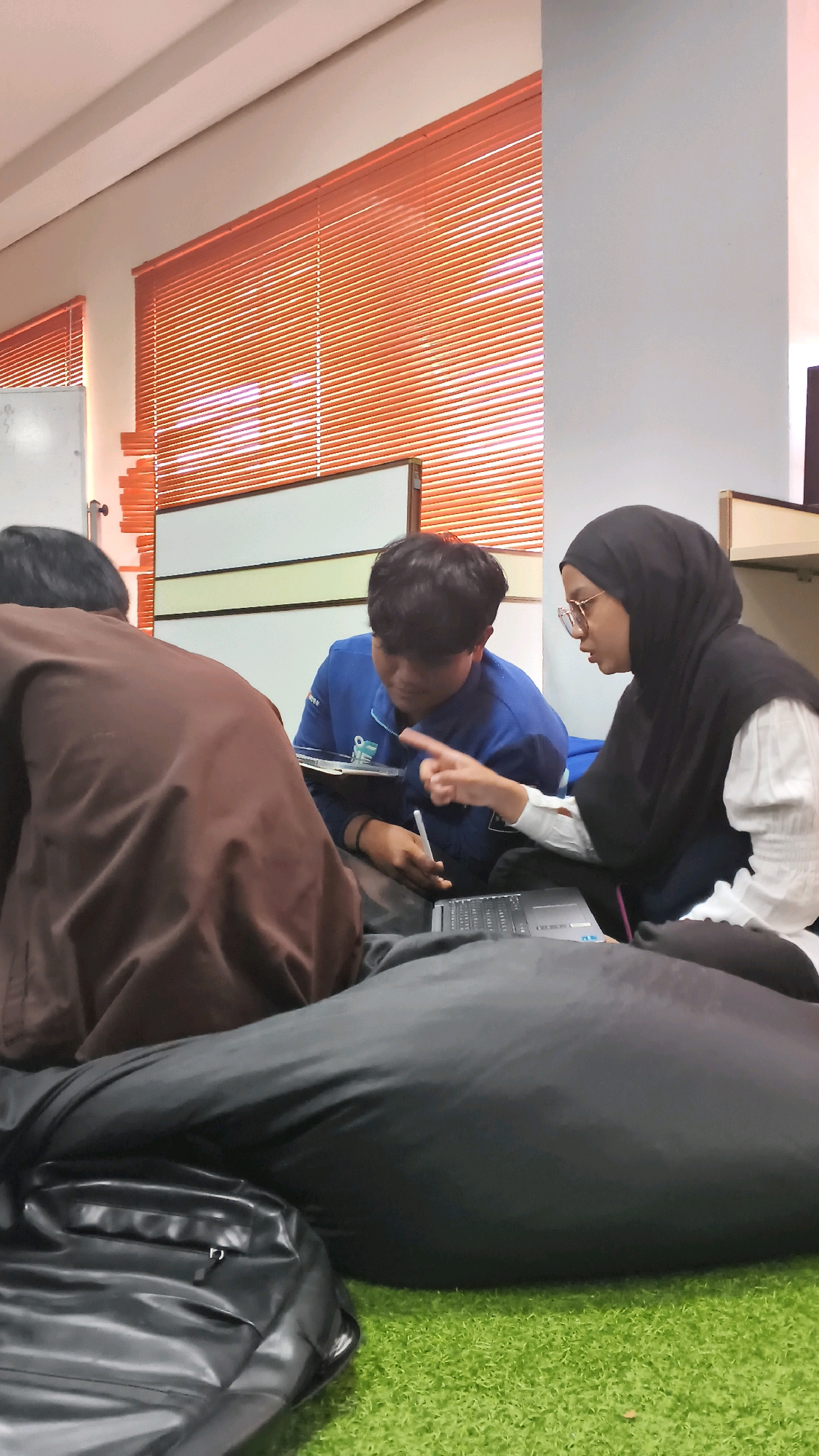 The image depicts three individuals in what appears to be a classroom or study space. In the foreground, a large, black beanbag chair is prominently displayed on a green, artificial turf surface. To the left, a person with dark hair is seated, their back partially to the camera, wearing a brown garment. In the center, a young male with dark hair is wearing a blue jacket and looking down at a tablet. To the right, a person wearing a black hijab and glasses is seated and gesturing with their right hand towards a laptop that is open on the beanbag. The background shows orange blinds covering a window and a white board. The lighting suggests an indoor environment, possibly during the daytime given the illumination from the window. The overall scene suggests a collaborative study session or a group project. The specific location in the city of Lohbener, Indonesia, is not discernible from the visual information provided in the image.