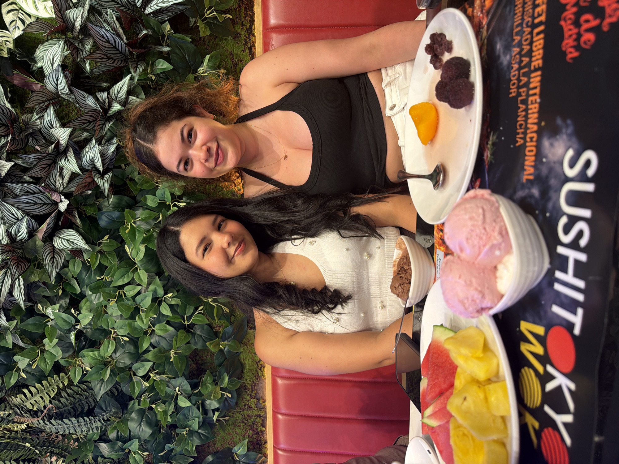 Two women are posing for a photo, seated in a restaurant booth with red leather upholstery. The woman on the left has wavy, reddish-brown hair and is wearing a black tank top and white shorts. She is smiling at the camera. The woman on the right has long, dark hair and is wearing a white top adorned with pearls. She is also smiling.
In front of them on a table are plates of food. One plate contains several small, dark berries and a slice of orange. Another plate holds fruit, including slices of watermelon and pineapple. There are also bowls of ice cream, one appearing to be strawberry flavored and the other chocolate. A menu with Spanish text is partially visible behind the food. The text on the menu includes "BUFFET LIBRE INTERNACIONAL", "RISCADA A LA PLANCHA", "RRILLA ASADOR", and "Sushi Wok".
The setting appears to be indoors, possibly a restaurant with a decorative wall of greenery behind the subjects. The lighting suggests it is daytime, but the exact time is not discernible. The overall mood is casual and happy, with both women appearing relaxed and enjoying their meal. The location context provided indicates the setting is Carretera De San Martín, Spain.