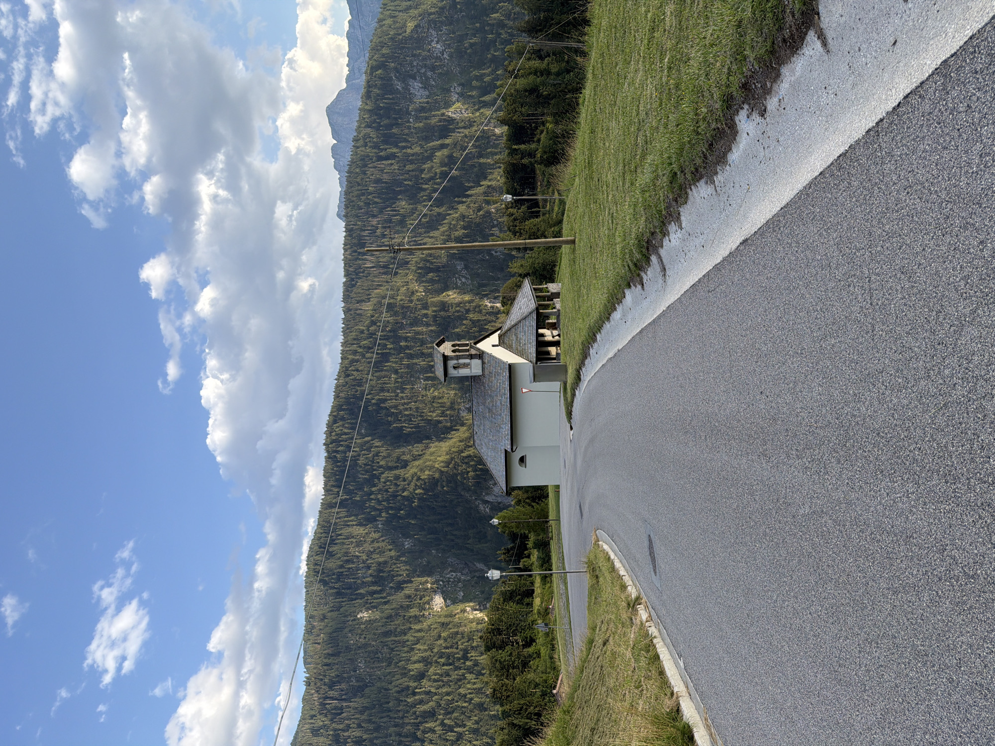 This is an eye-level, medium shot of a winding asphalt road leading towards a small, light-colored building with a gray roof, possibly a chapel, situated on a grassy hill. The building has an arched entrance porch and a small bell tower. It is nestled against a backdrop of a dense, dark green forest that covers a steep mountainside. In the distance, rugged, grey mountain peaks are visible against a bright blue sky with scattered white clouds. A wooden utility pole with wires stands to the right of the building. The road is bordered by a grassy verge on the left and a mowed grassy area on the right, which slopes gently upwards towards the building. The lighting suggests it is daytime, likely late afternoon due to the shadows. The location is identified as Sauris / Zahre, Italy, a village known for its unique dialect and traditions, nestled in the Carnic Alps.